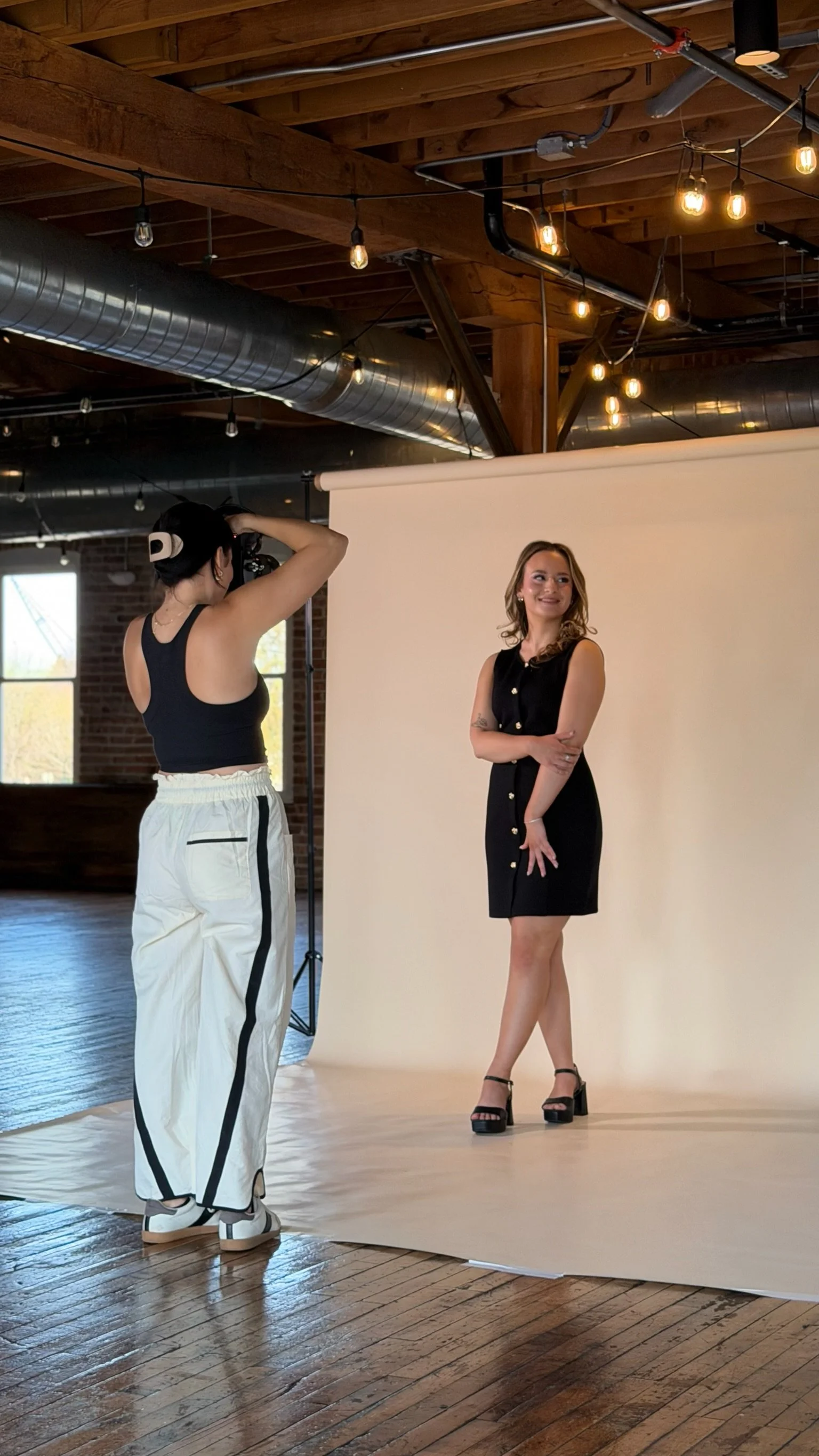A woman in a black sleeveless dress and black high-heeled sandals posing for a photo in front of a white backdrop, with a photographer taking her picture indoors under warm lighting.