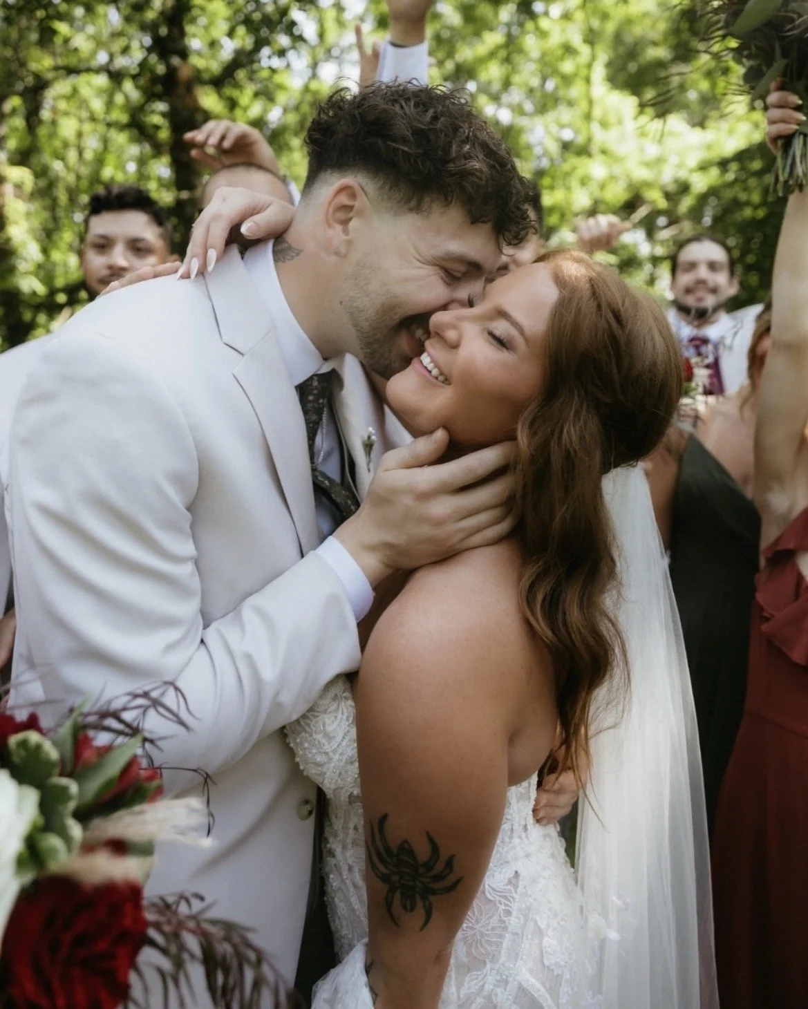 A happy couple on their wedding day, embracing each other outdoors with green trees in the background, surrounded by friends and family celebrating.
