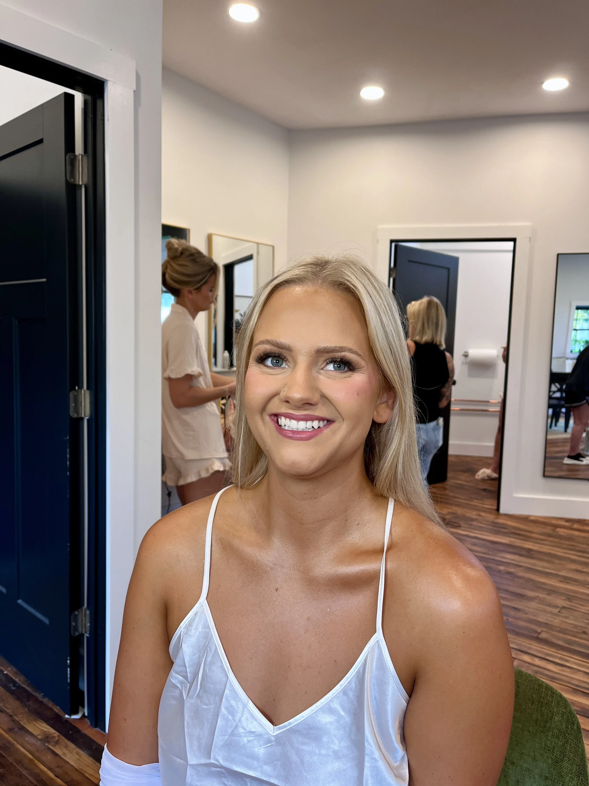A smiling woman with blonde hair wearing a white satin dress, sitting in a room with white walls and wooden flooring, with three other women in the background