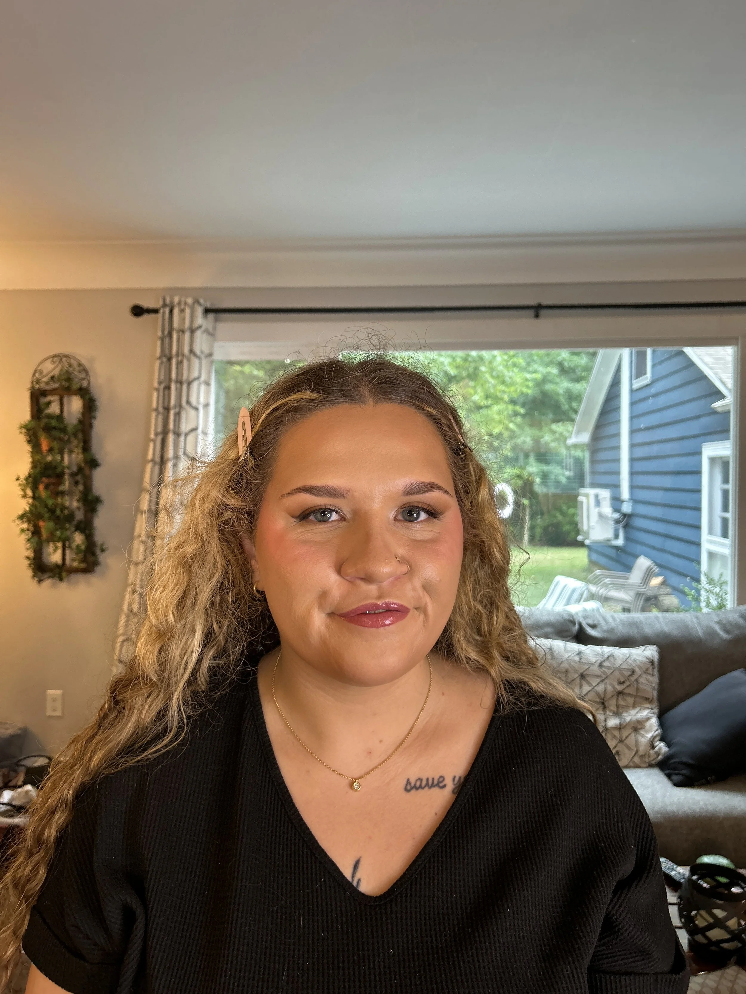 A woman with long curly hair and a tattoo on her chest, wearing a black top and a necklace, sitting in a living room with a window and house outside in the background.