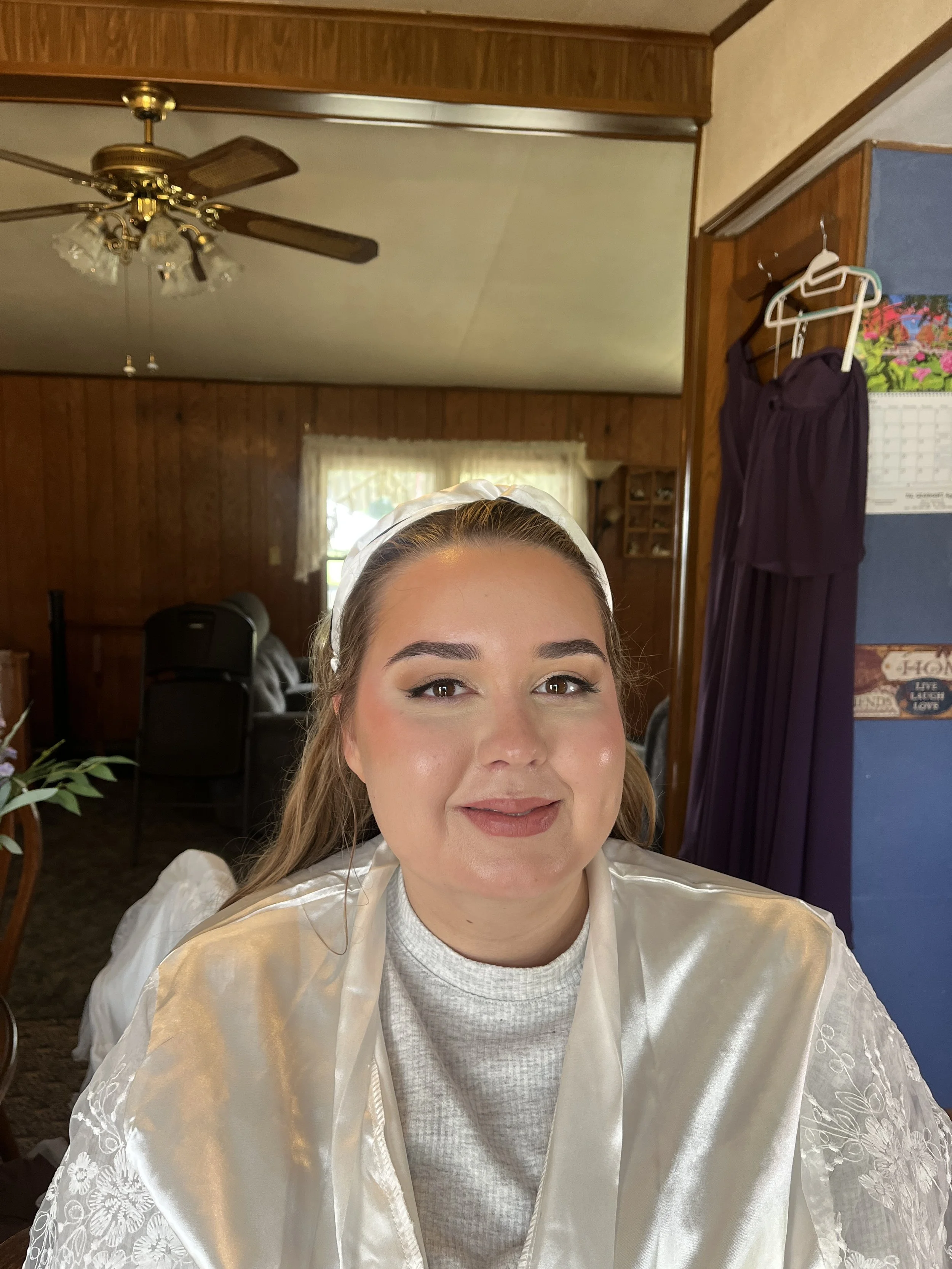 A woman with styled brown hair and makeup, wearing a white satin robe and a white headband, smiling at the camera in a wooden-paneled room with a ceiling fan and decorated walls.