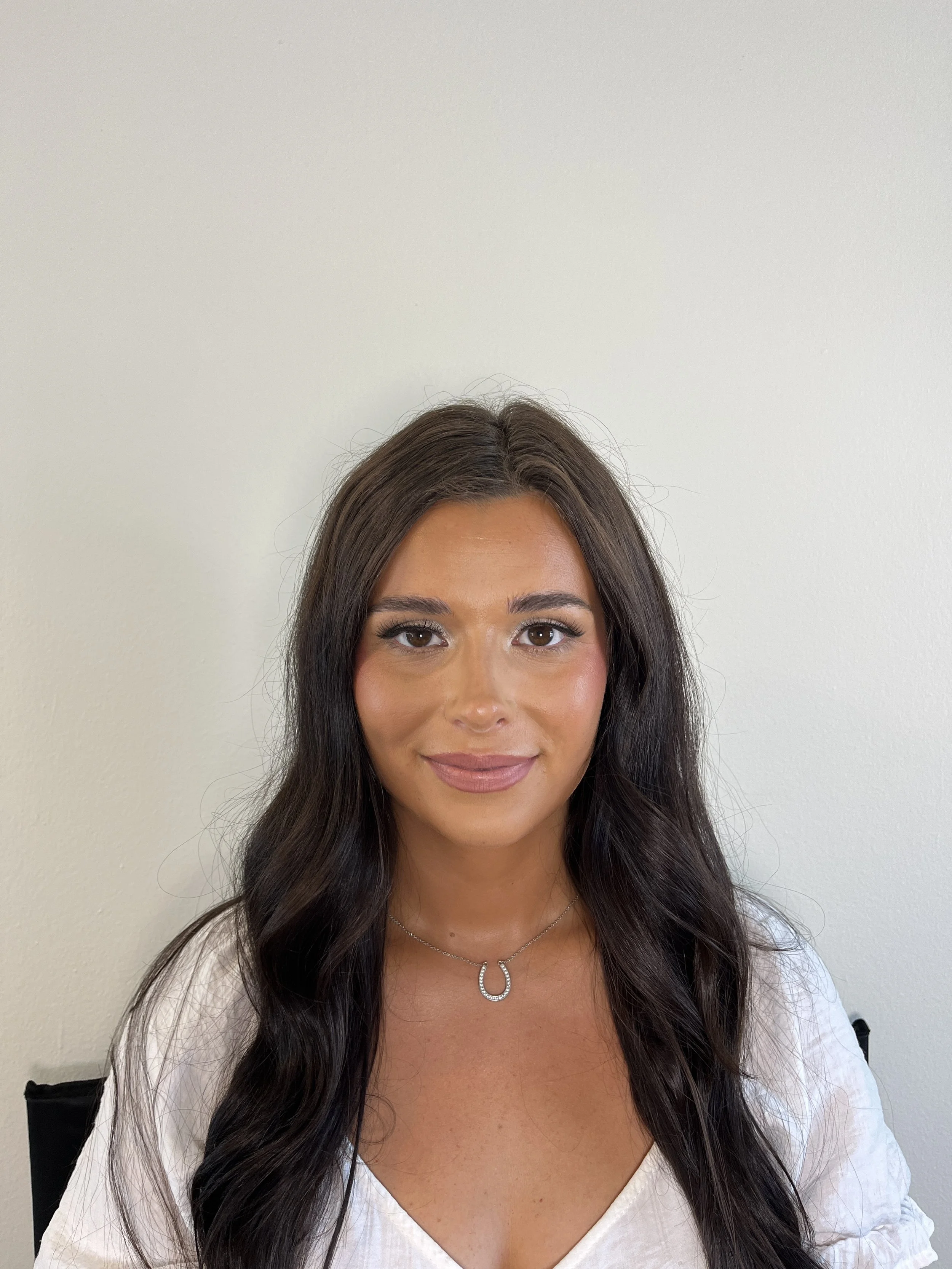 Close-up of a woman with long dark hair, wearing a white top, a silver horseshoe necklace, and subtle makeup, standing against a plain off-white wall.