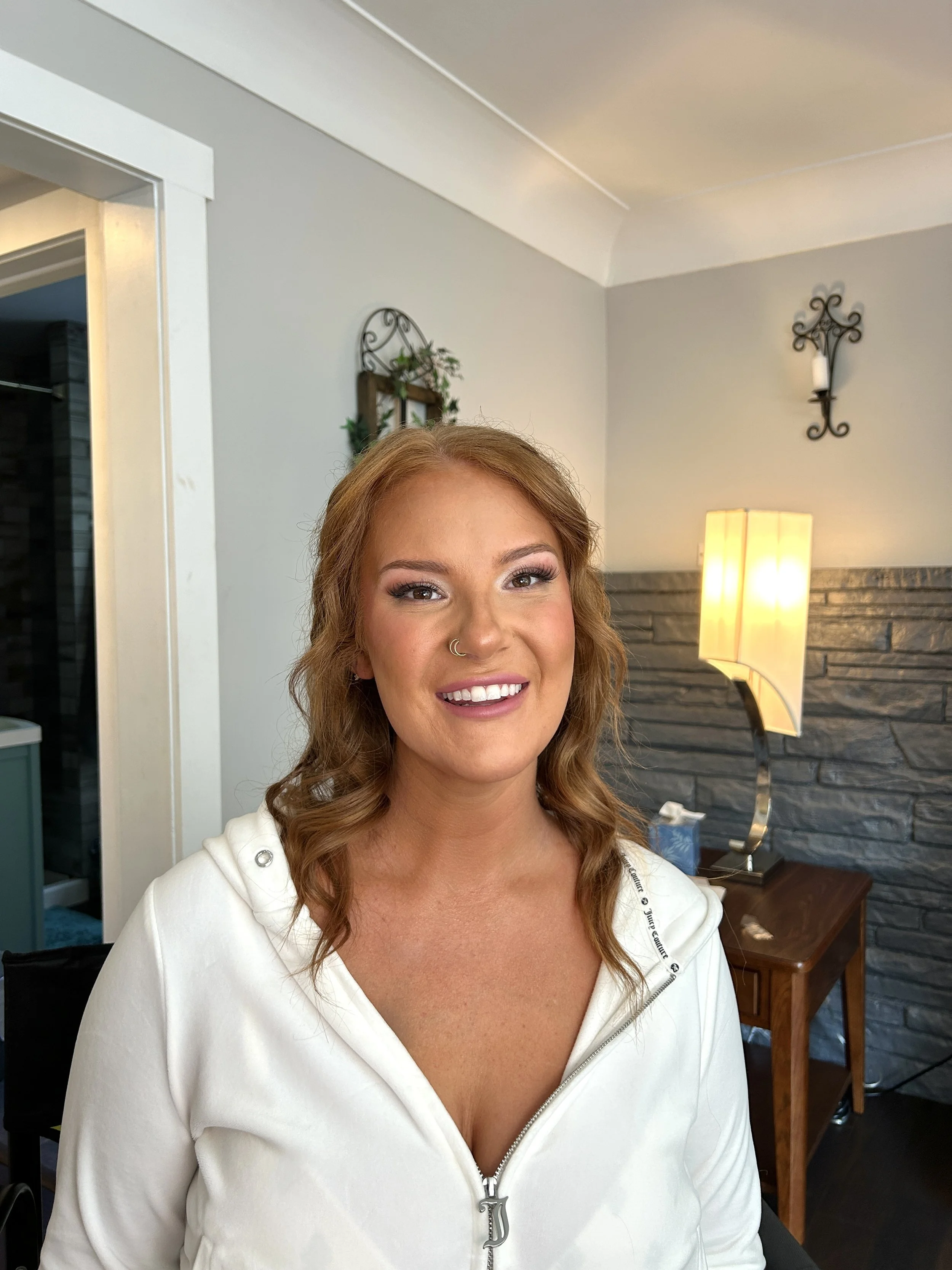 Smiling woman with red curly hair, nose ring, and white hoodie, indoors with modern decor and a lamp in the background.
