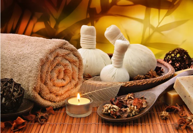 Spa scene with rolled towel, white herbal compress balls, a lit candle, herbal leaves, and soap on a bamboo mat with warm lighting.