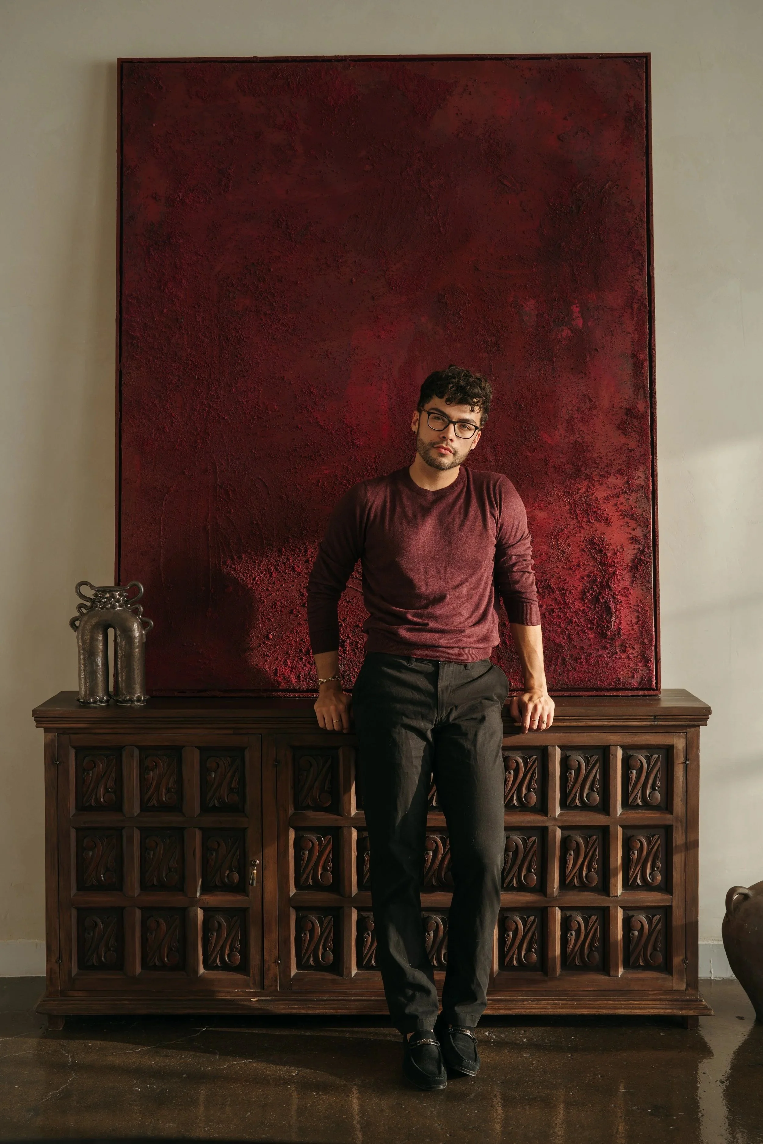 A man with curly dark hair, glasses, and a beard is leaning against a wooden cabinet with intricate carvings, in front of a large red textured artwork on a beige wall. He is wearing a maroon long-sleeve shirt and black pants.