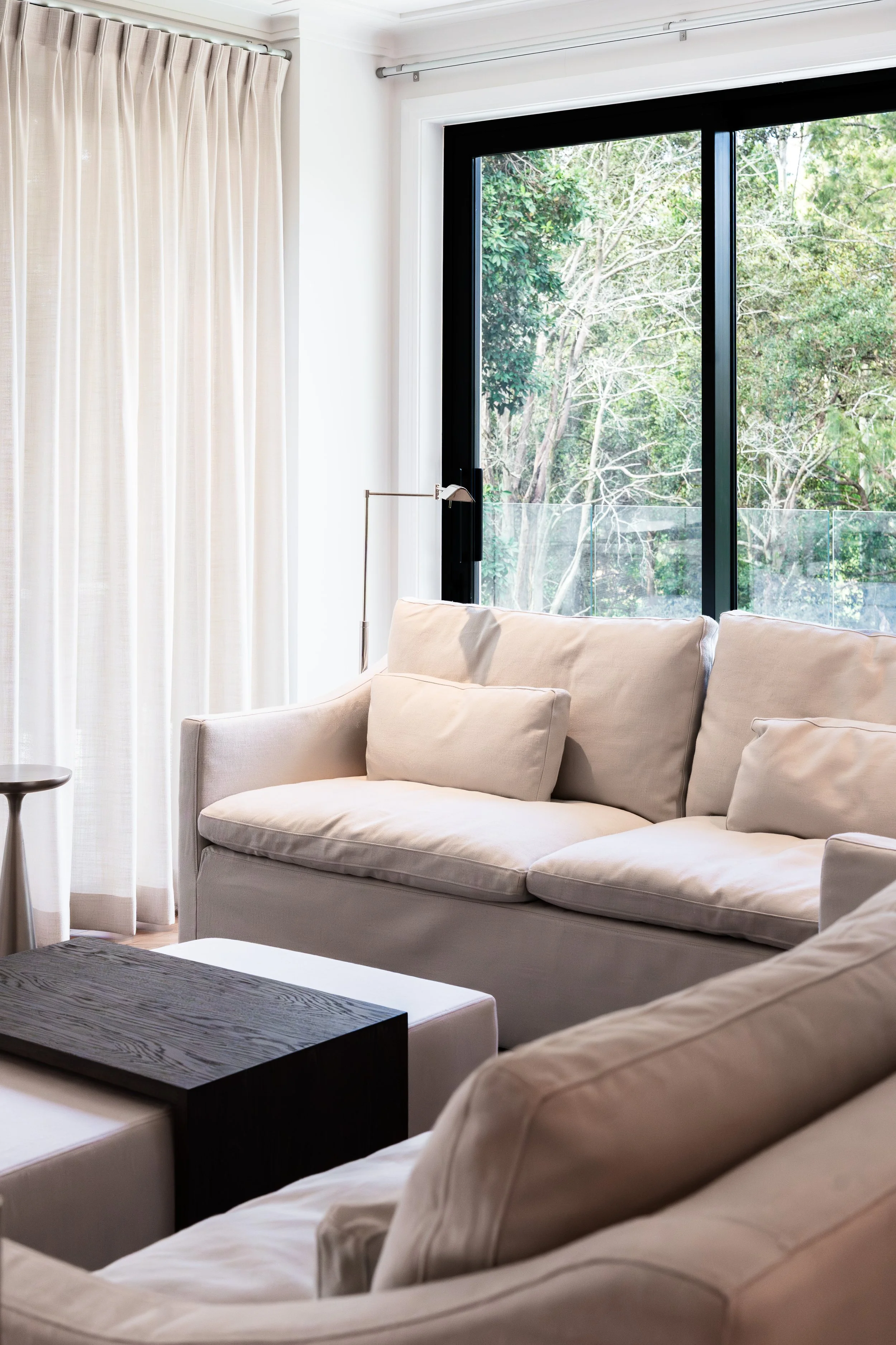 Living room with beige sofa, black-framed window showing trees outside, and white curtains.