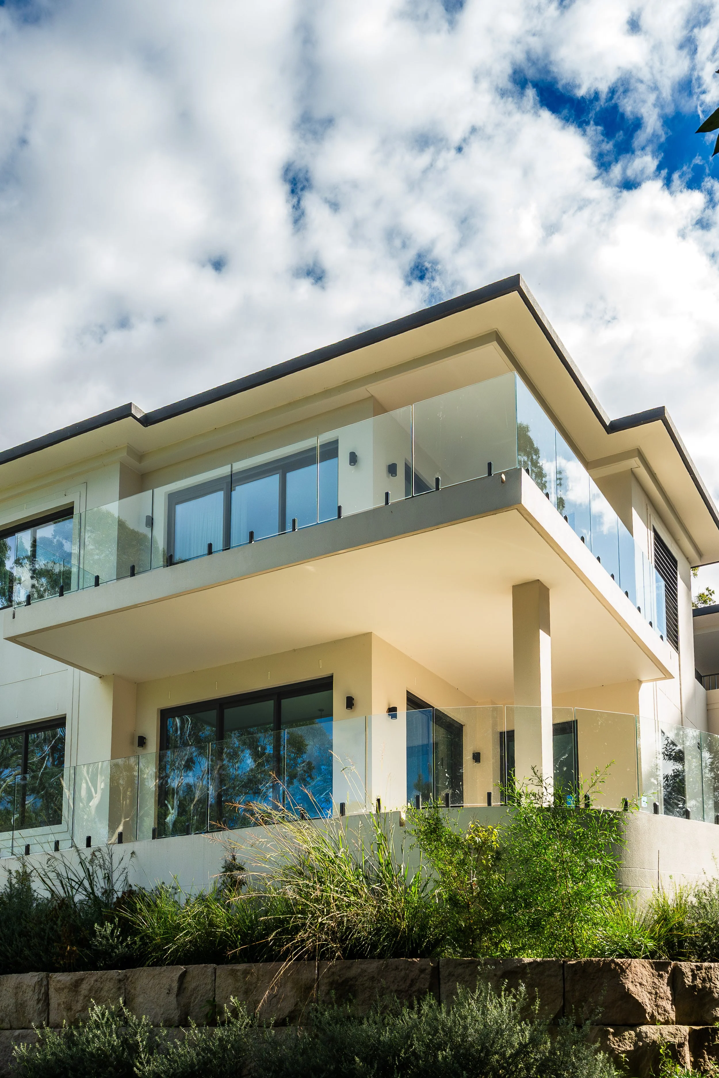 Modern building with glass balconies, large windows, and a flat roof, set against a cloudy sky and surrounded by greenery.
