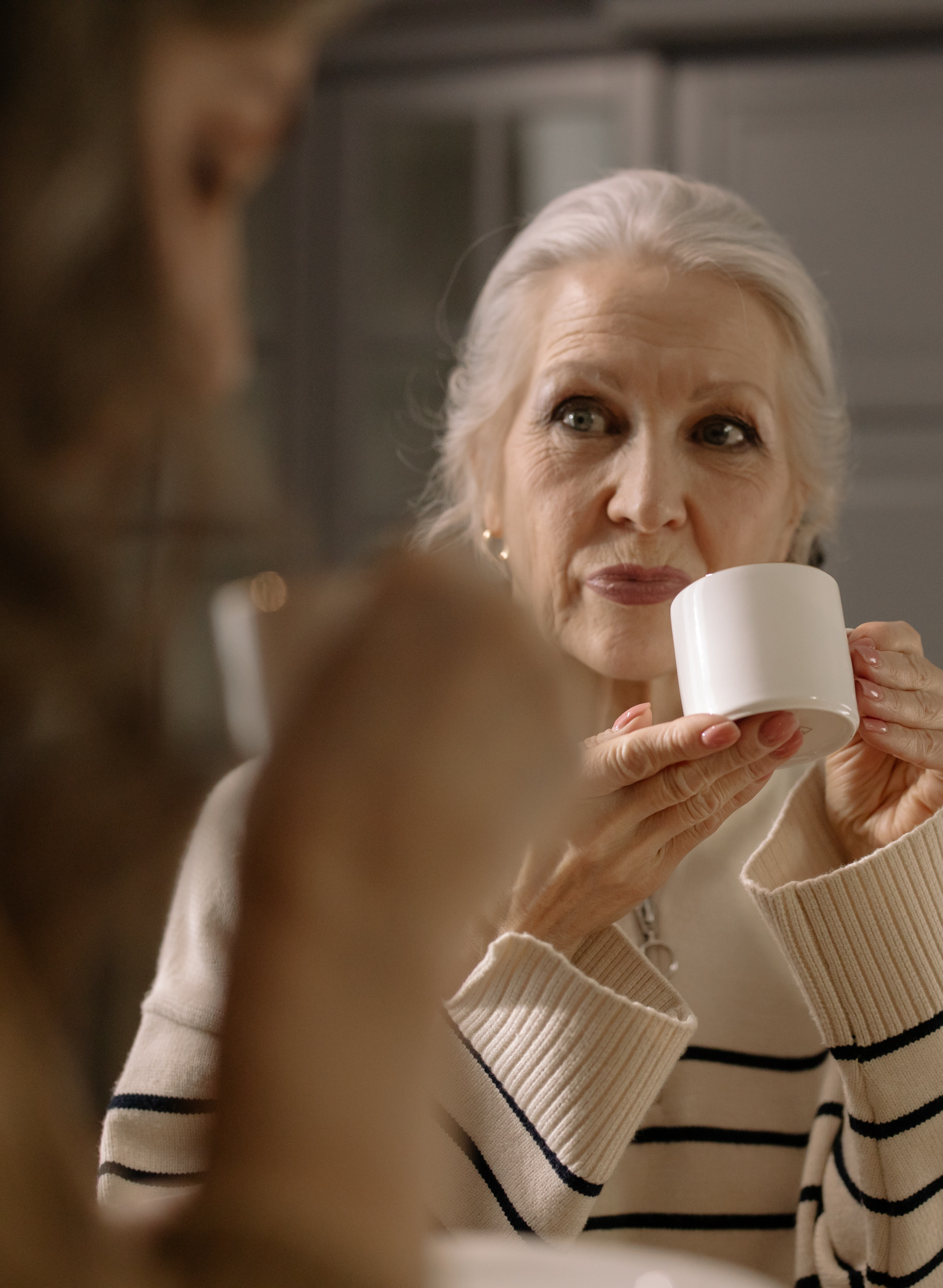 An elderly woman with silver hair sipping from a white mug while sitting at a table, having a conversation with a person whose face is partially visible in the foreground.