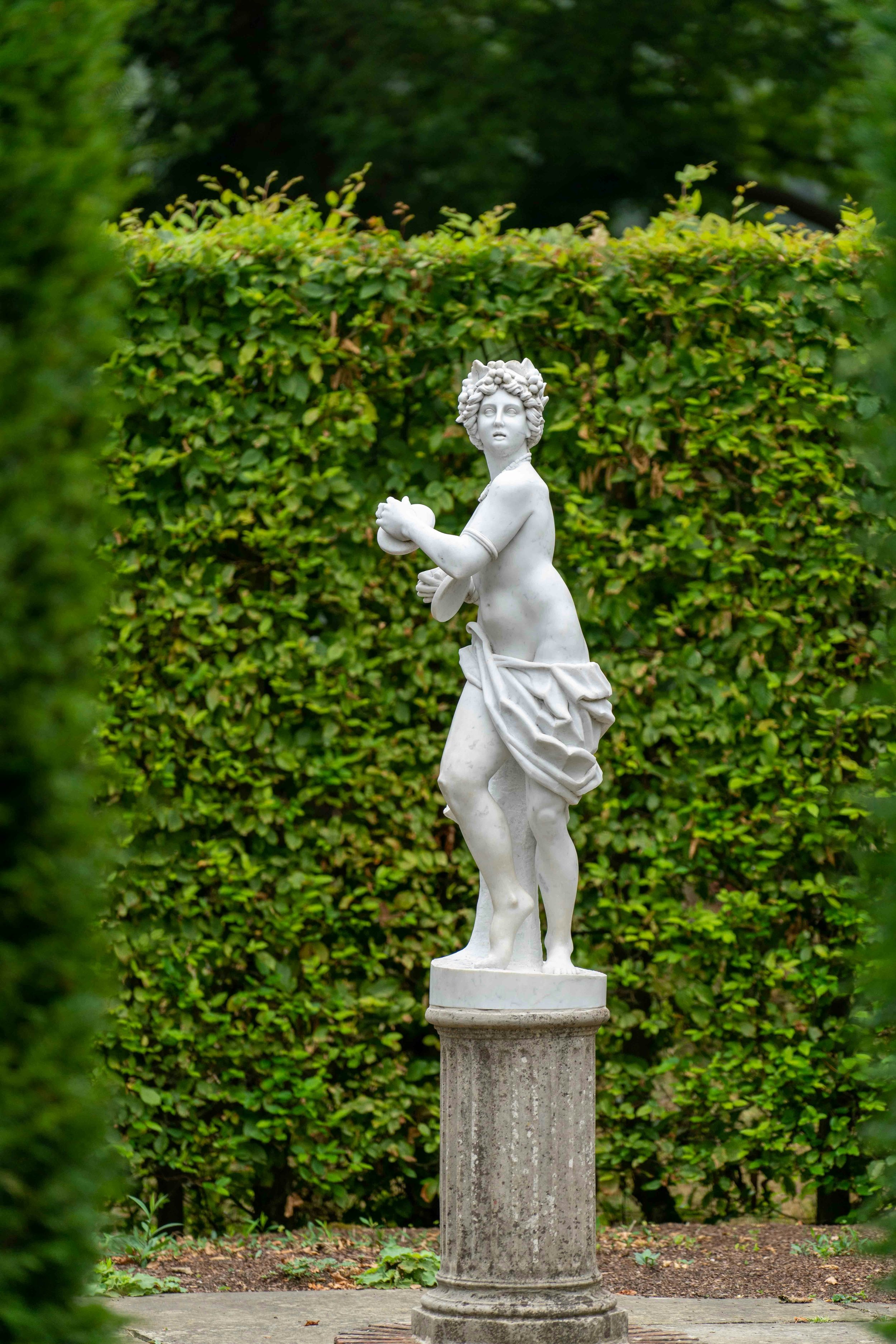 A classical white marble statue of a young woman standing outdoors on a stone pedestal, surrounded by green bushes.
