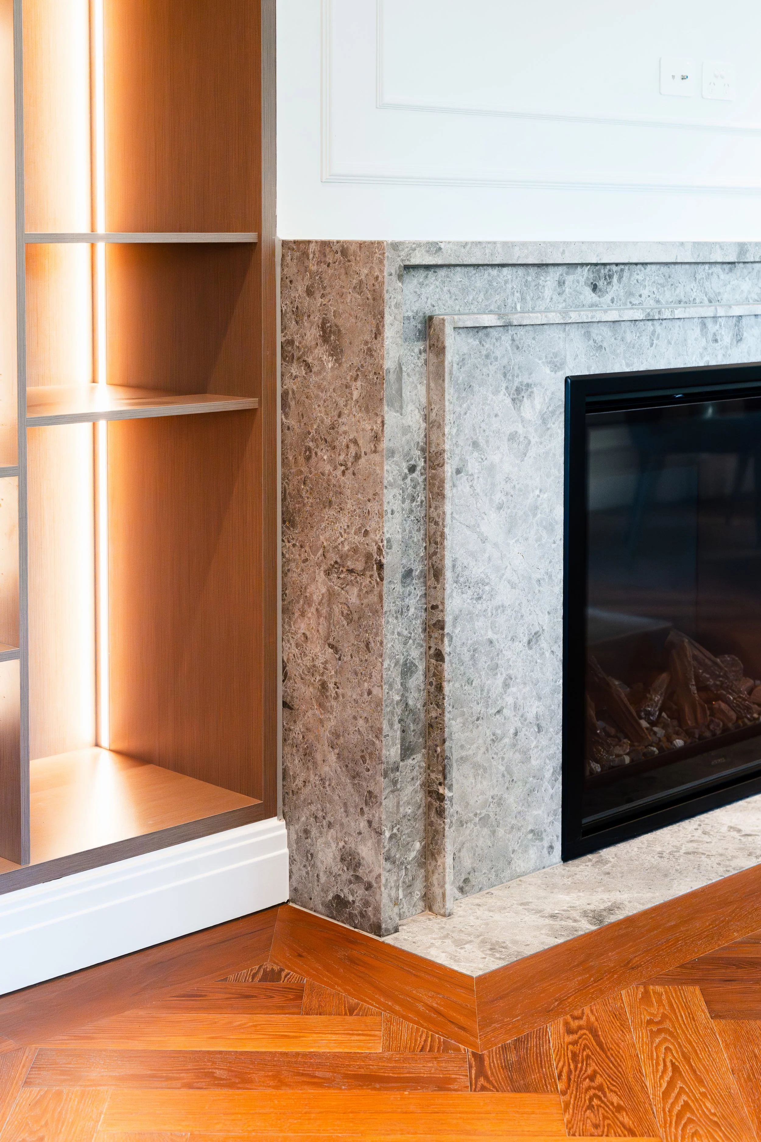 Close-up of a contemporary fireplace with a grey marble surround, a white wall with electrical outlets, and a wooden floor with a herringbone pattern.