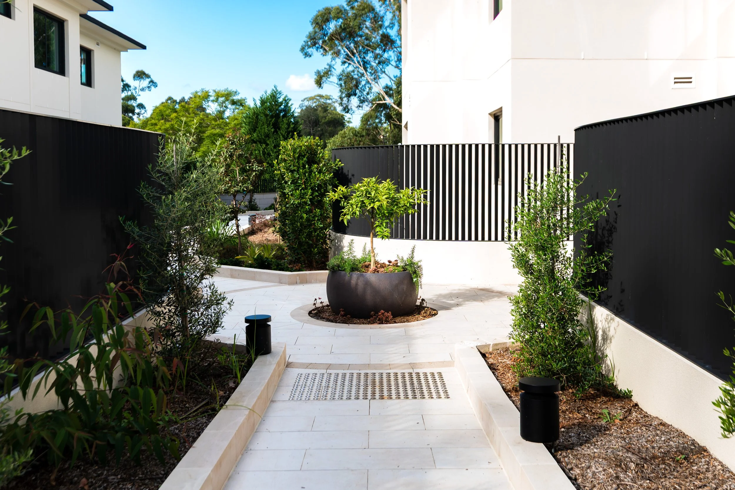 Modern outdoor patio area with white tiled flooring, black fencing, potted plants, and surrounding greenery.
