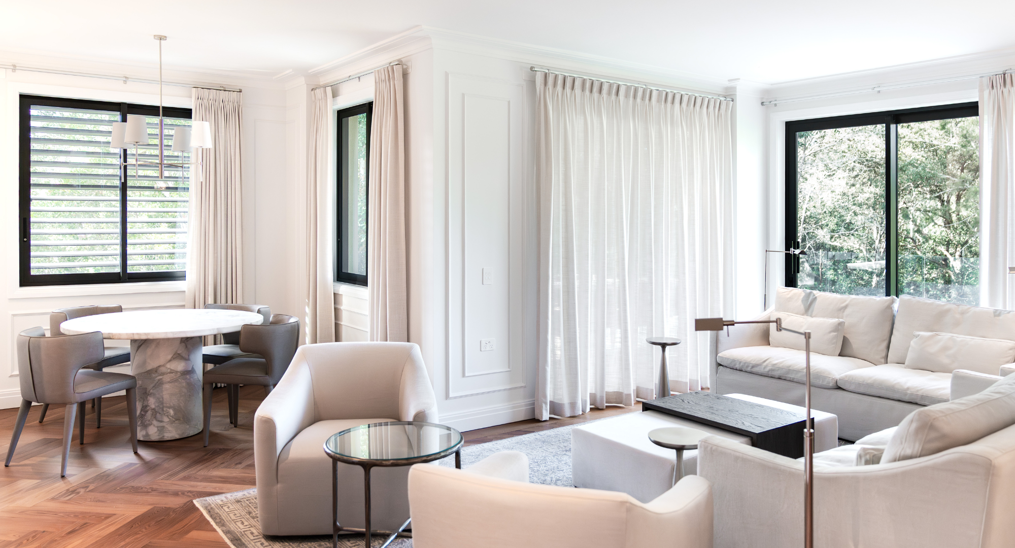 Modern living room with white sofa, armchair, round dining table, and large windows with curtains, showcasing greenery outside.