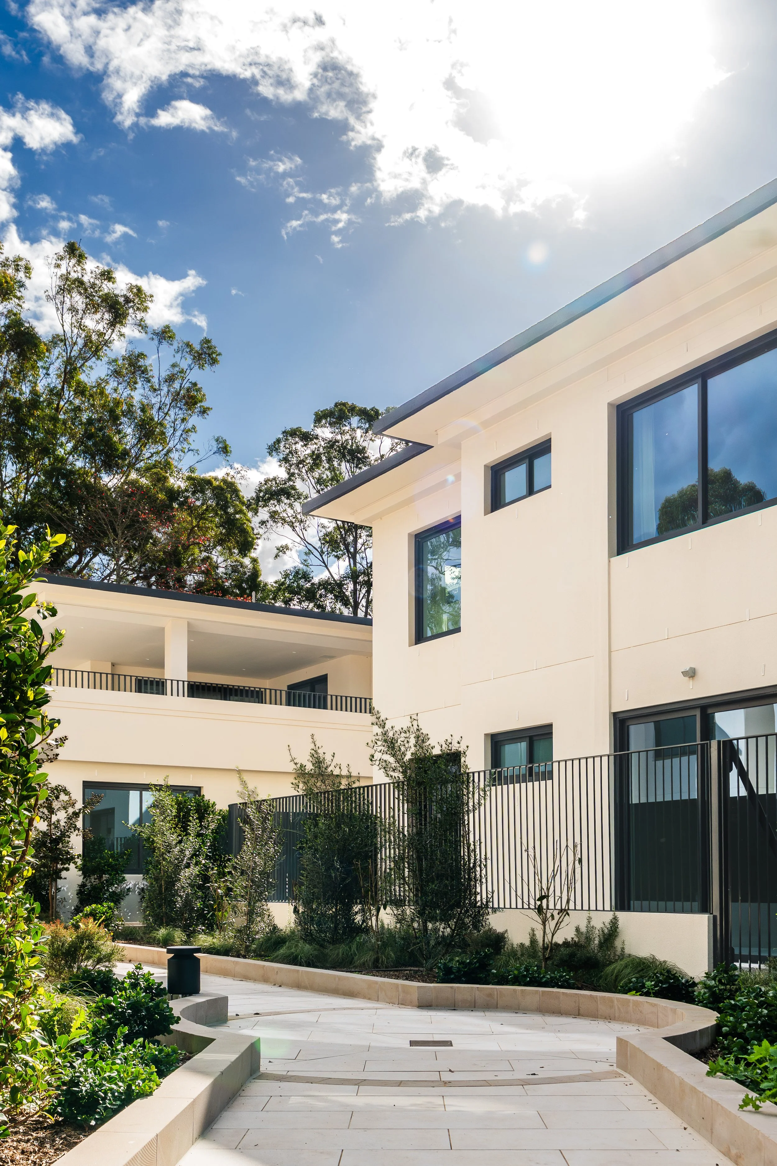 Exterior view of a modern multi-story residential building with large windows, set against a backdrop of a partly cloudy sky and tall trees, with a landscaped pathway and garden area in the foreground.