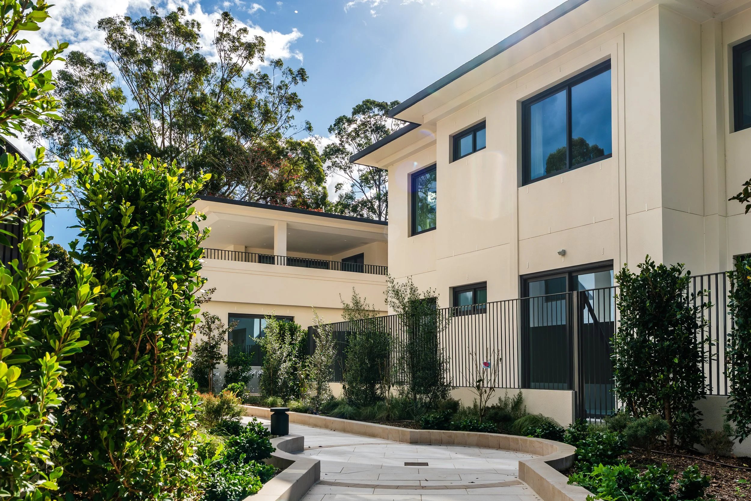 Modern multi-story white residential building with large black-framed windows and balcony railings, surrounded by well-maintained garden with various small plants and bushes, in a bright sunny day with a blue sky and some clouds.