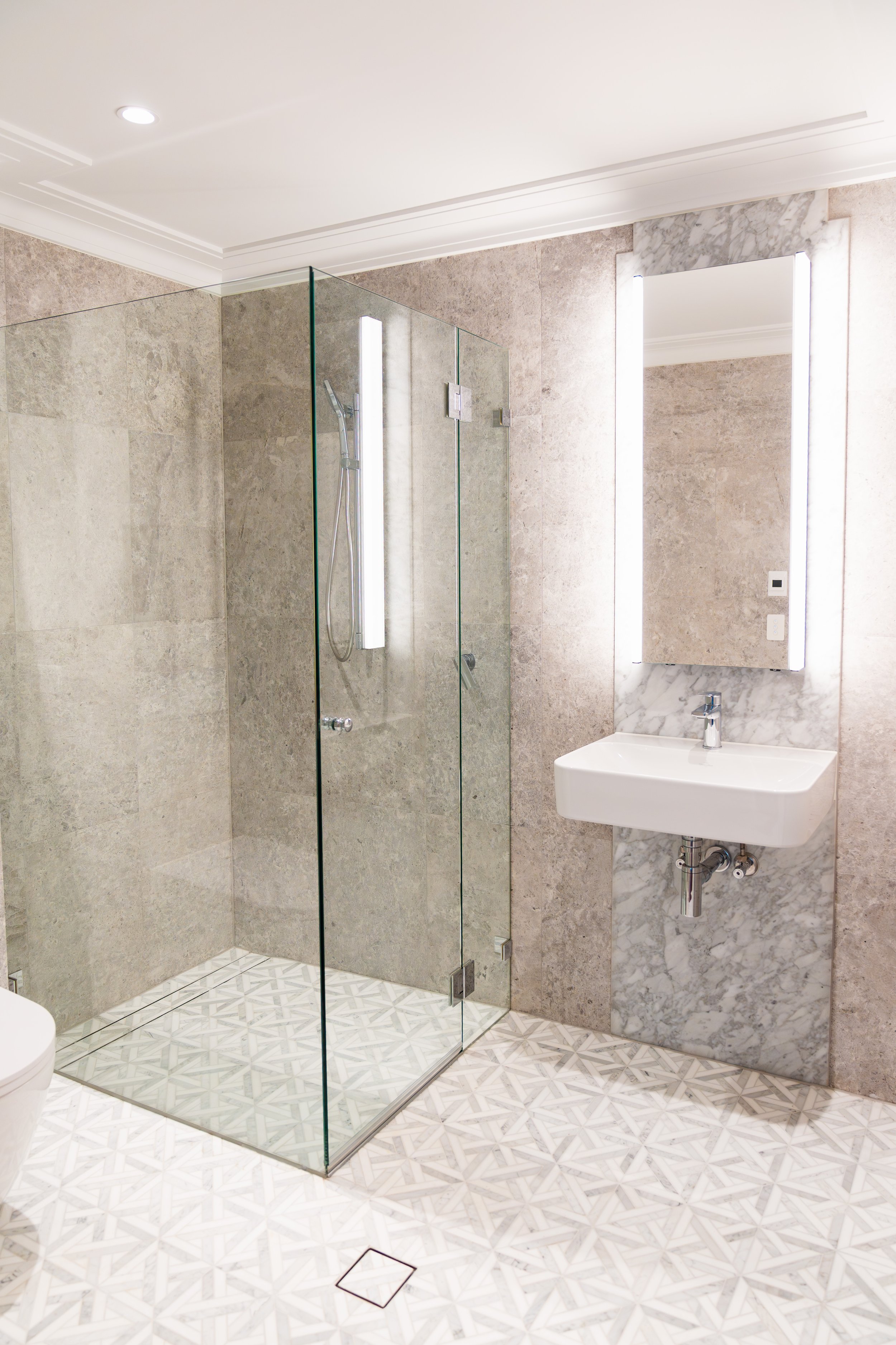 Modern bathroom with a walk-in shower enclosed by glass, beige tile walls, a white rectangular sink, and a large mirror with integrated lighting. The floor has patterned tiles, and there is a marble accent on the wall behind the sink.