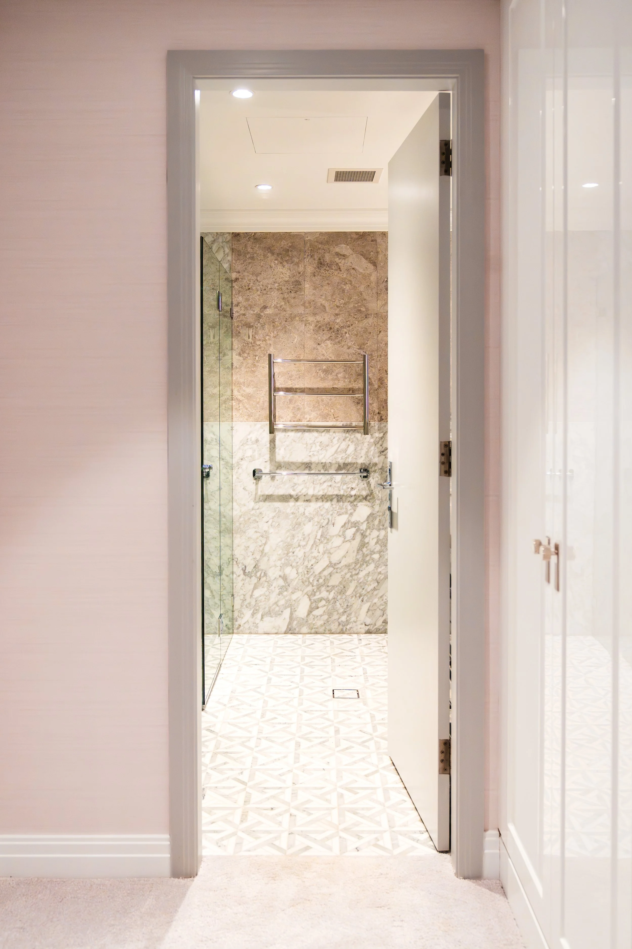 View into a bathroom through an open door, showing a marble shower with a glass door, a towel rack, and beige tiled walls, with patterned flooring.