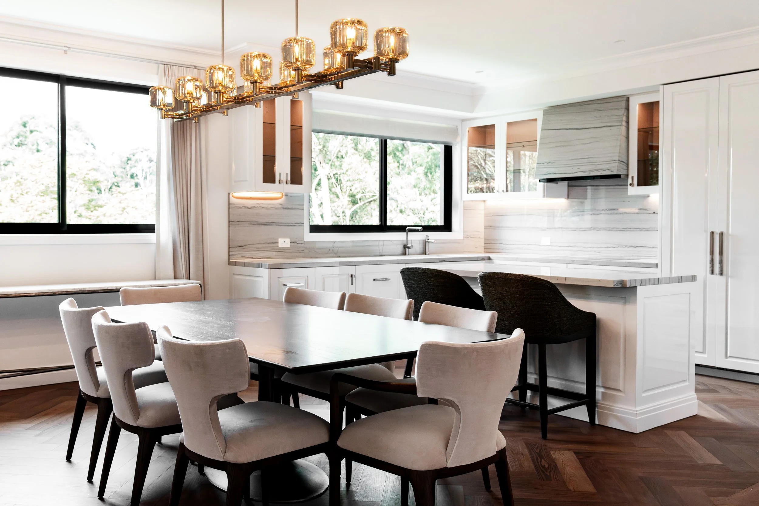 Modern kitchen with white cabinets, marble backsplash, large windows, and a dining table with beige chairs. A gold chandelier hangs from the ceiling.