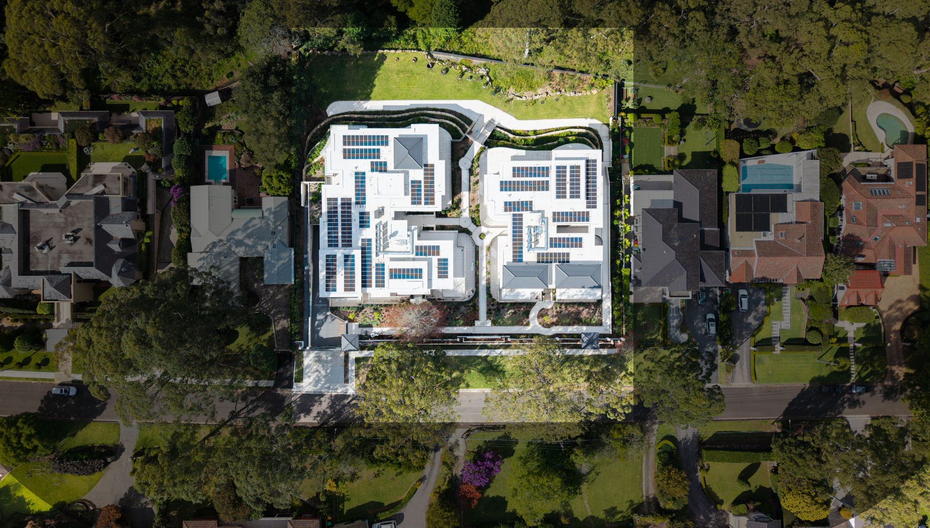 Aerial view of a large residential property with a modern house featuring solar panels on the roof, surrounded by greenery, trees, and neighboring houses with swimming pools and well-maintained lawns.