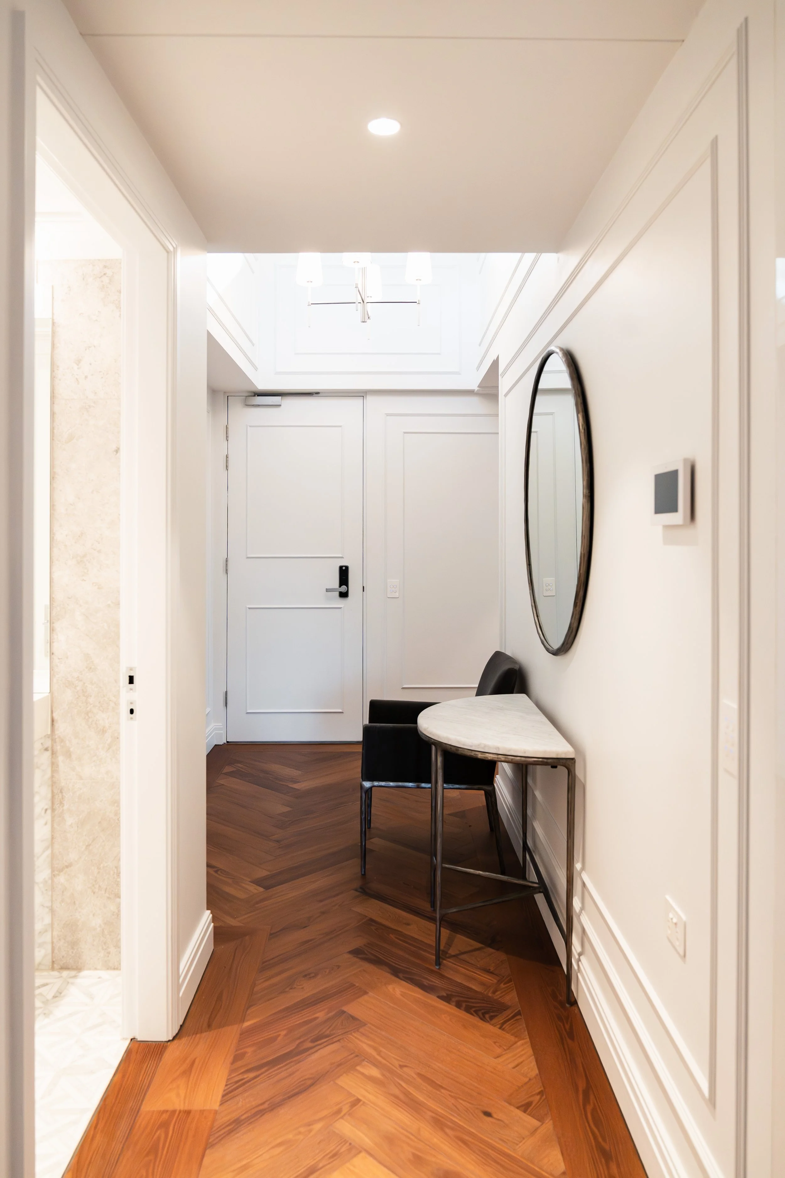 Interior hallway with white walls, a round mirror, a small marble-topped table, and black chairs. The floor has a herringbone wood pattern, and there is a closed white door at the end of the hallway.