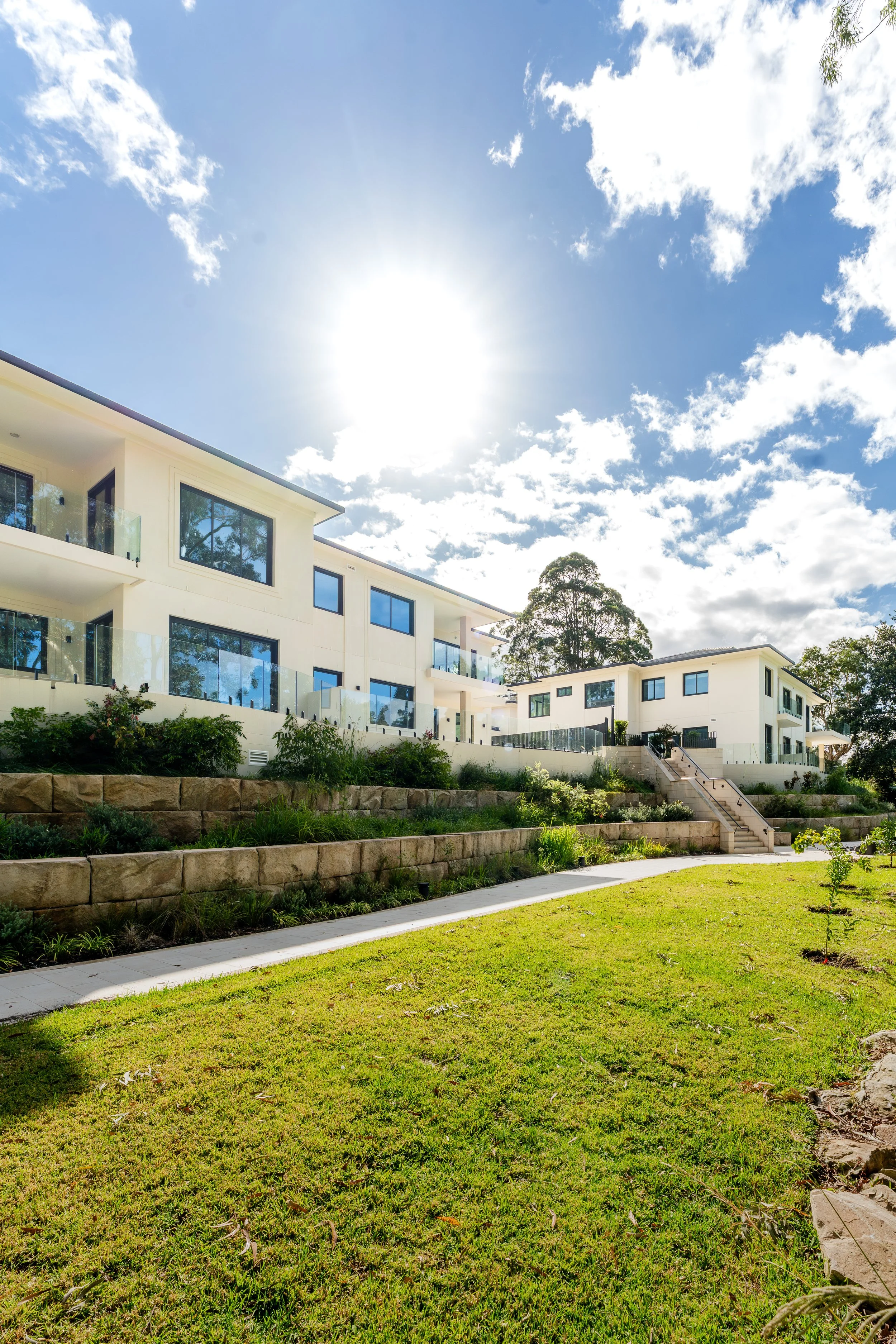 Modern multi-story white residential buildings with large windows and glass balconies, surrounded by landscaped garden with a grassy lawn, trees, and stone retaining walls, under a partly cloudy sky with the sun shining brightly.