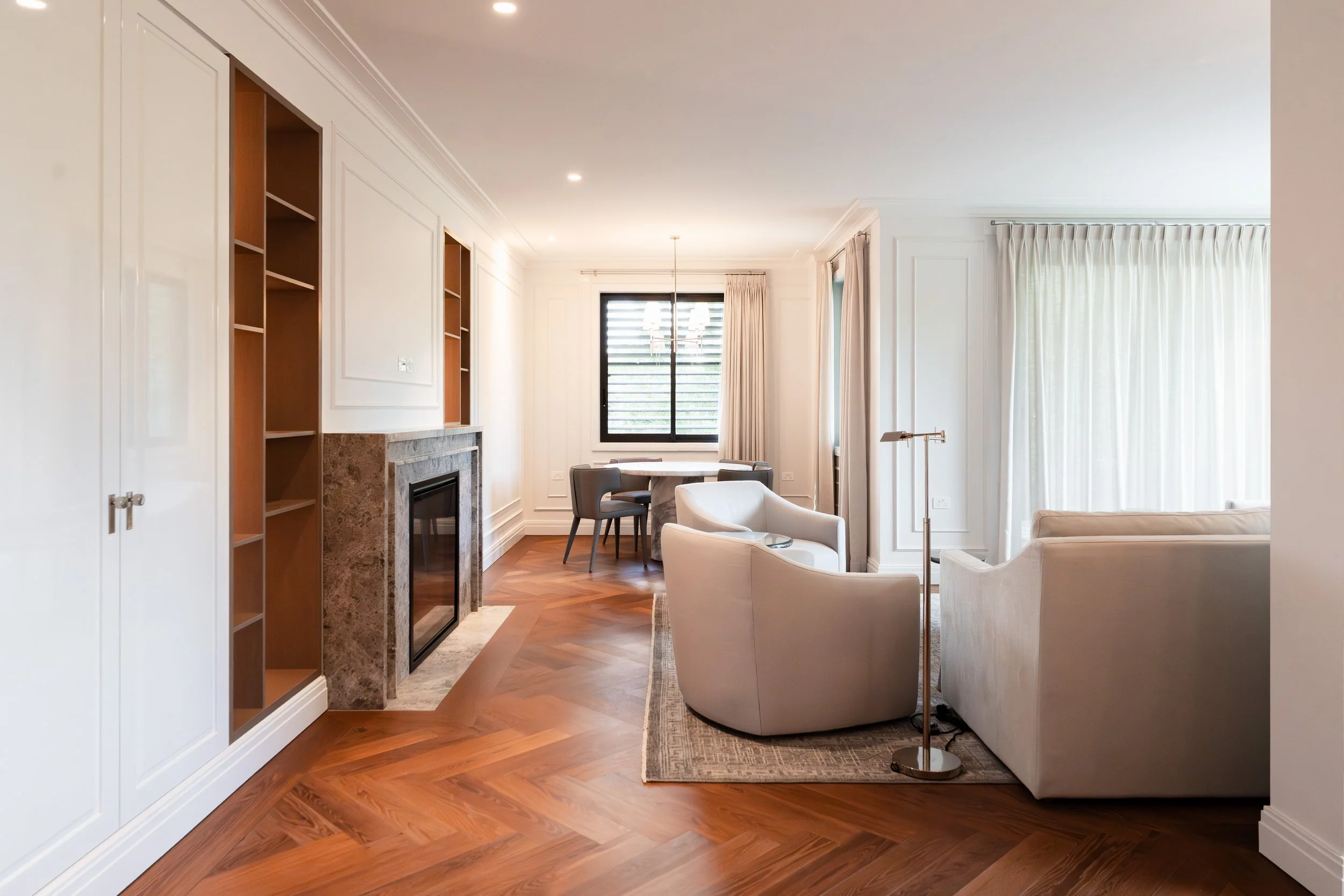 A spacious living room with white walls, hardwood floors, and large windows with curtains. The room features a fireplace with a marble surround, a round dining table with chairs, an armchair, a sofa, and a floor lamp, all arranged to create a cozy an