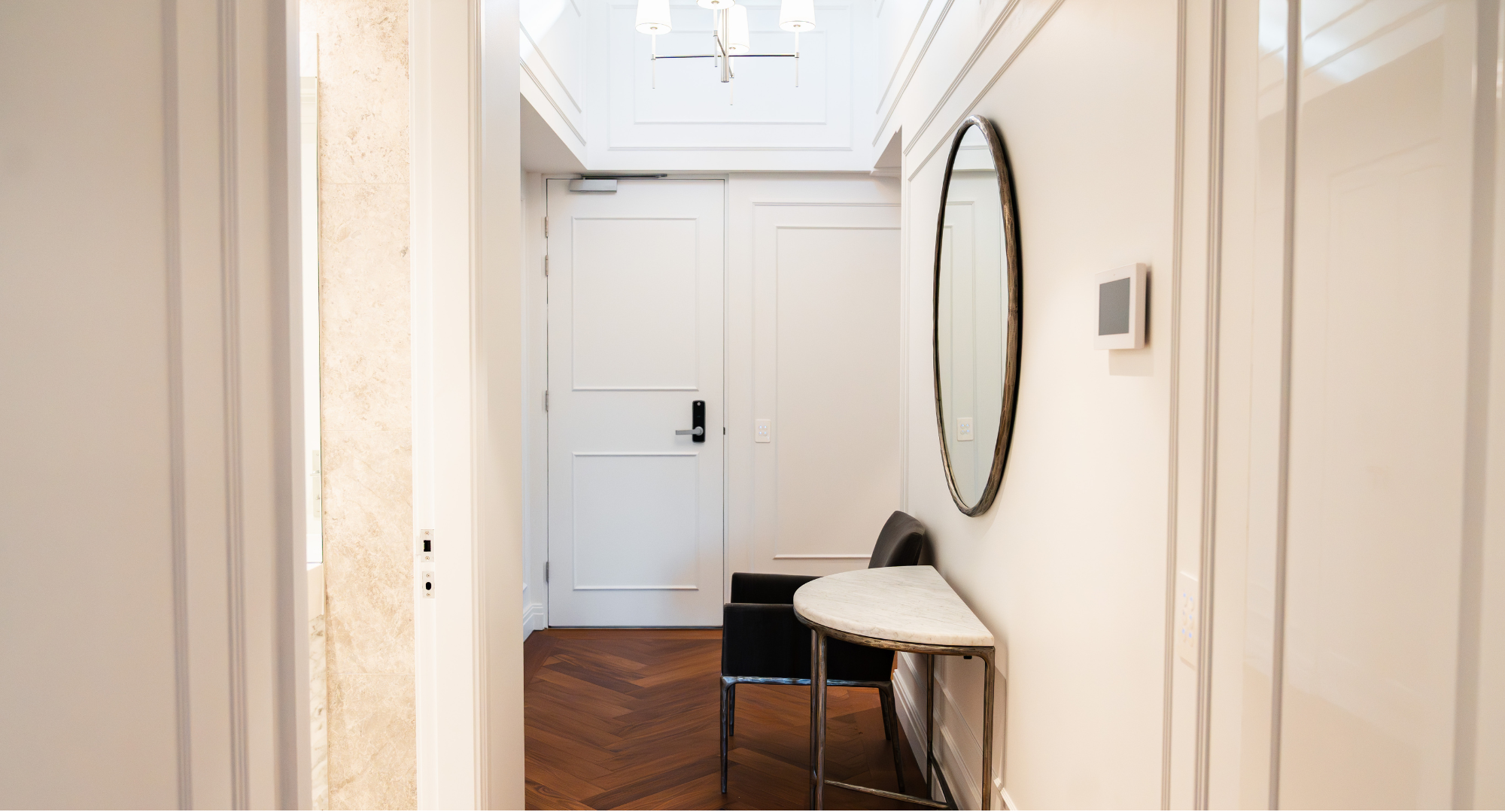 Interior hallway with white walls, a door, a round mirror, a small table, and black chairs.