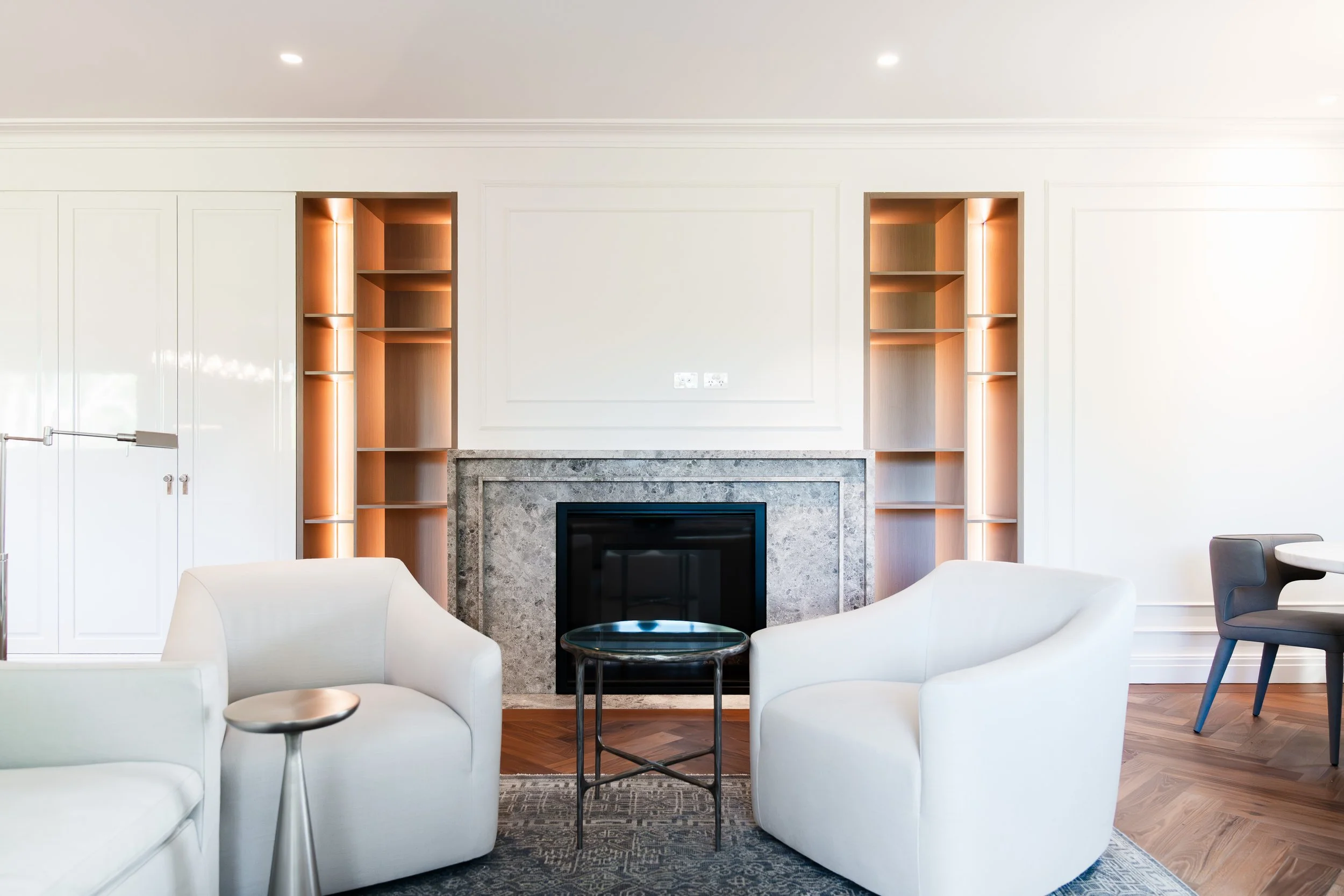 Modern living room with white chairs, a marble fireplace, and built-in wooden shelves with warm lighting.