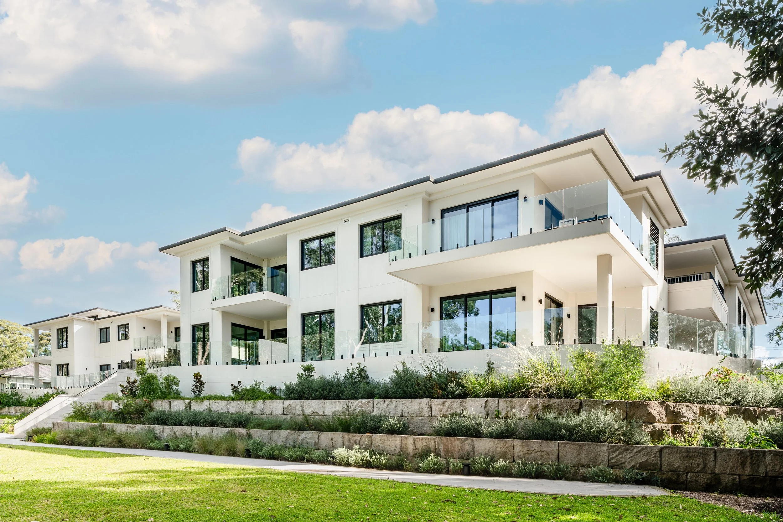 Modern white multi-story apartment building with glass balconies, large windows, surrounded by greenery under a partly cloudy sky.