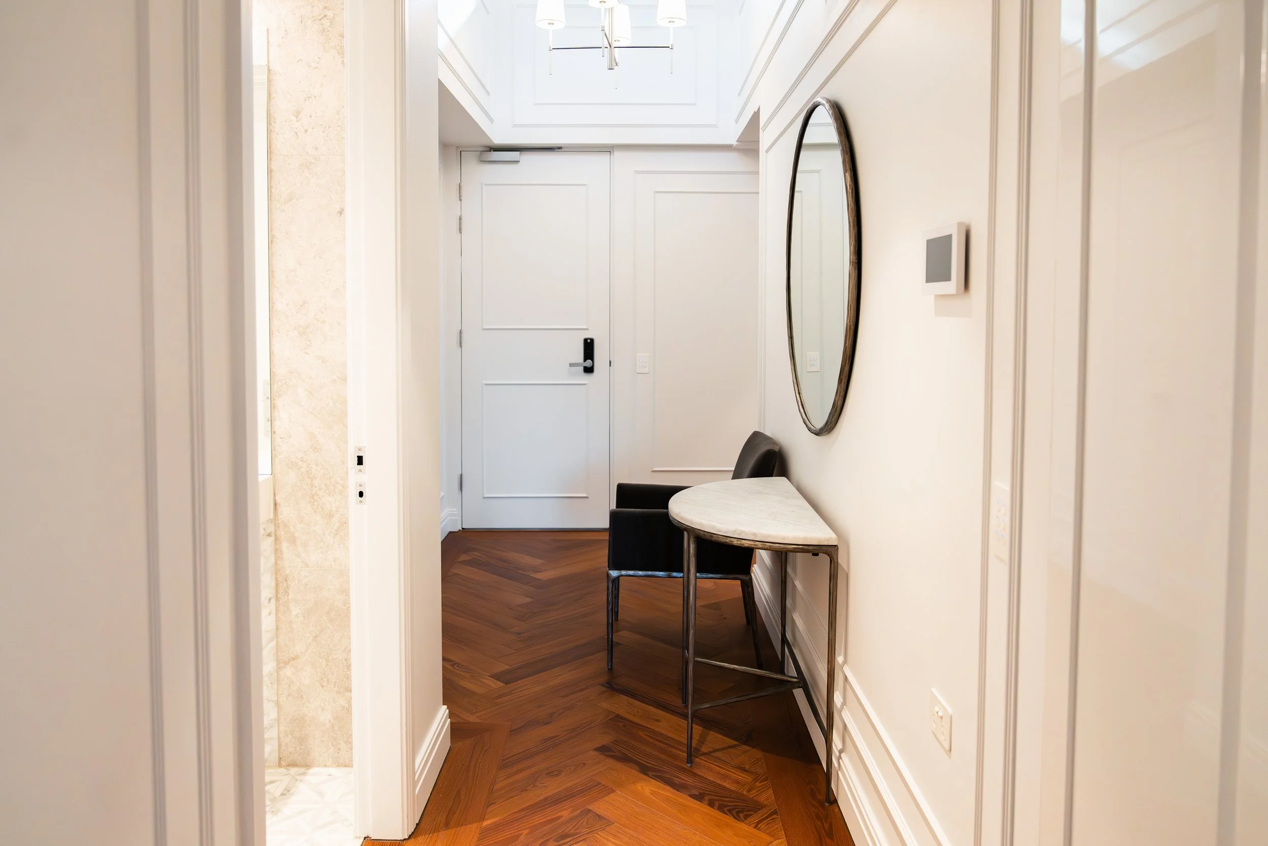 Hallway with white walls and trim, a round table with two black chairs, a large oval mirror on the wall, and a white door at the end with a modern light fixture hanging from the ceiling.