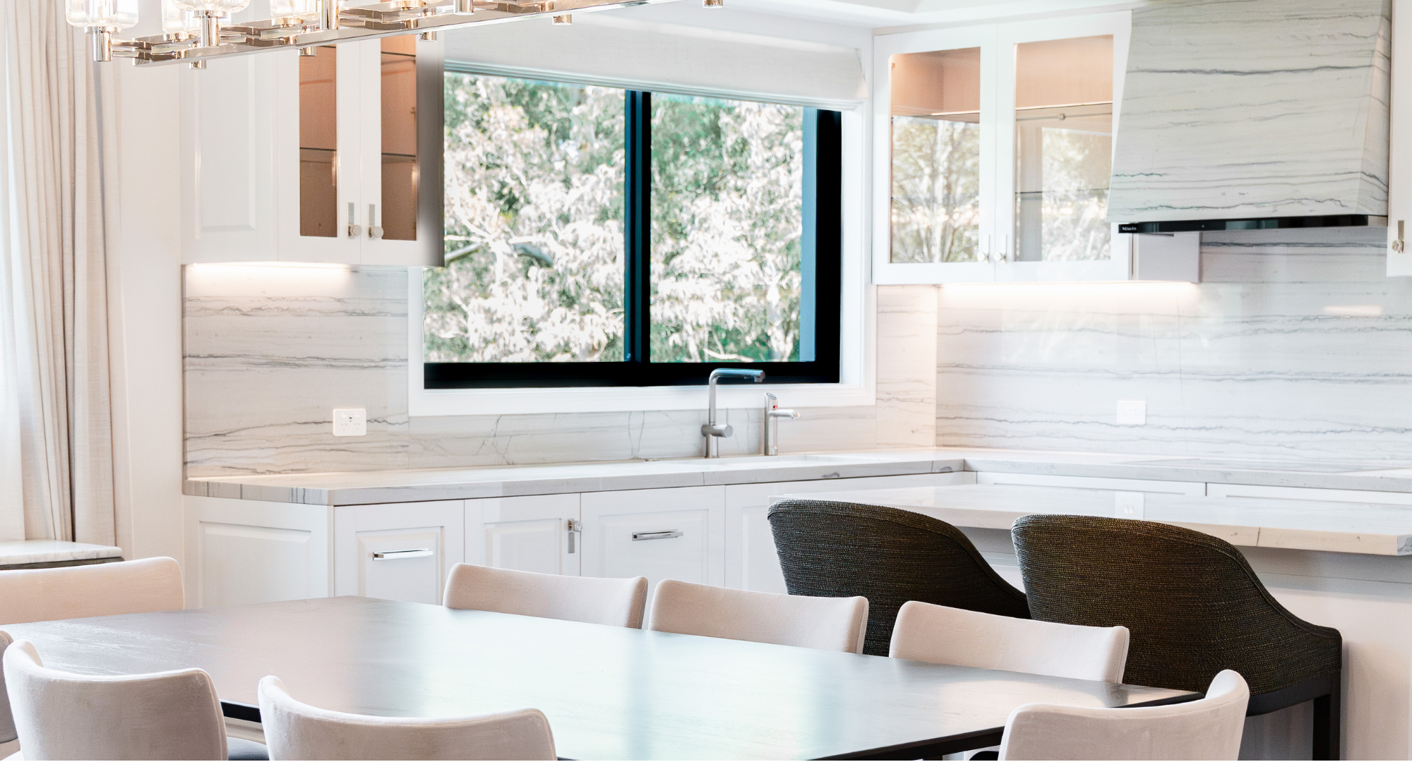 Modern kitchen with white cabinets, marble backsplash, black window, and dining table with beige and dark gray chairs.