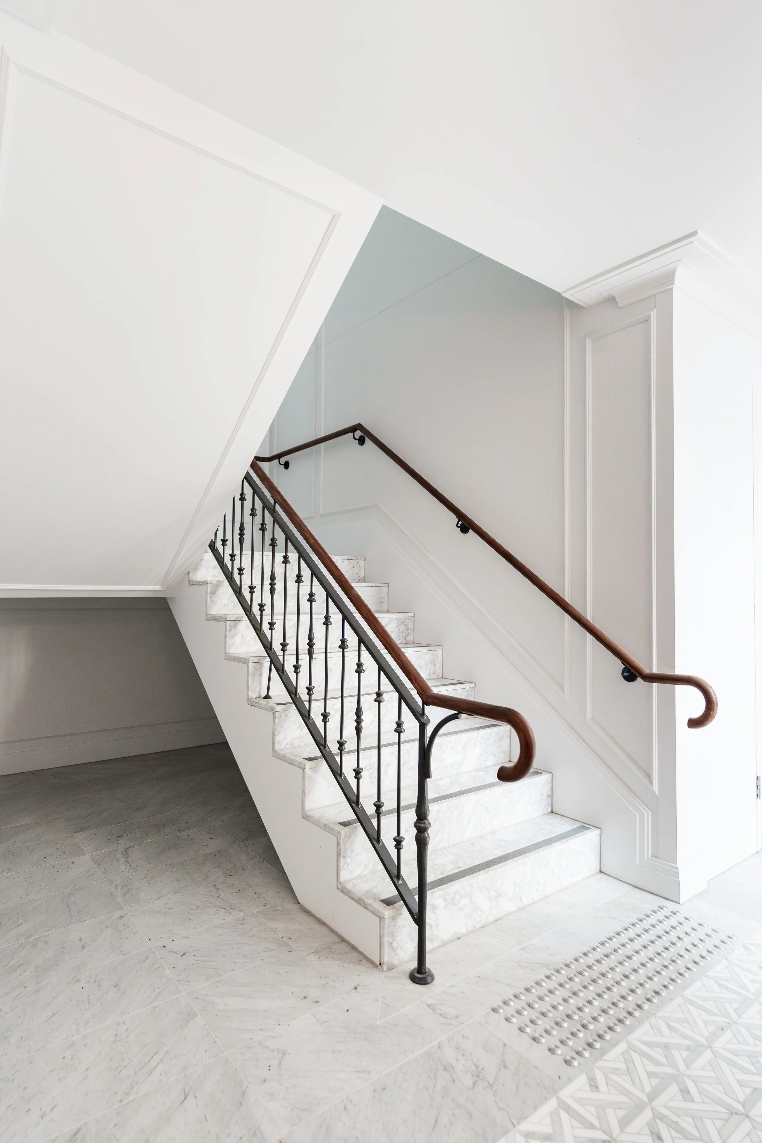 Interior view of a staircase with marble steps, black metal railing with wooden handrail, white walls, decorative wall panel, and flooring with marble tiles, near a vent grille.