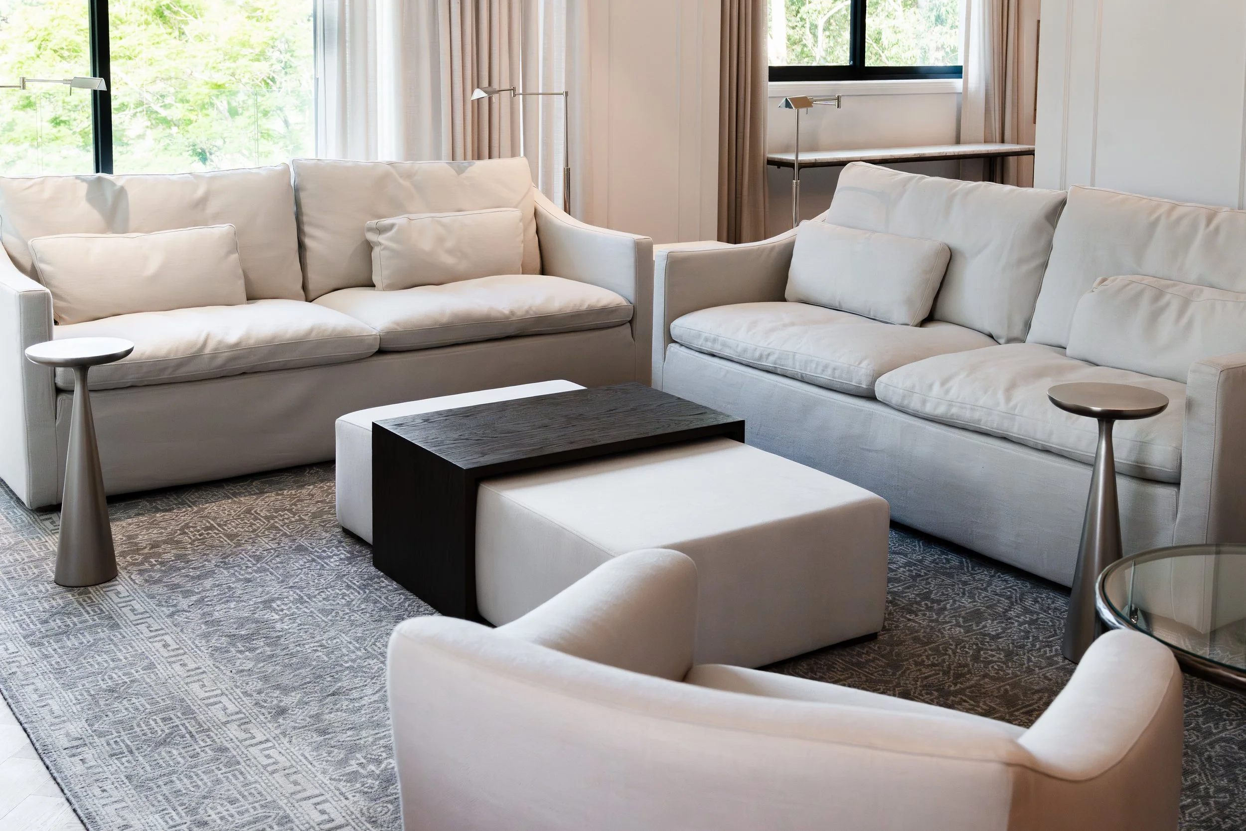 A modern living room with white sofas, a black coffee table, and round side tables, large window with curtains, and a view of green trees outside.