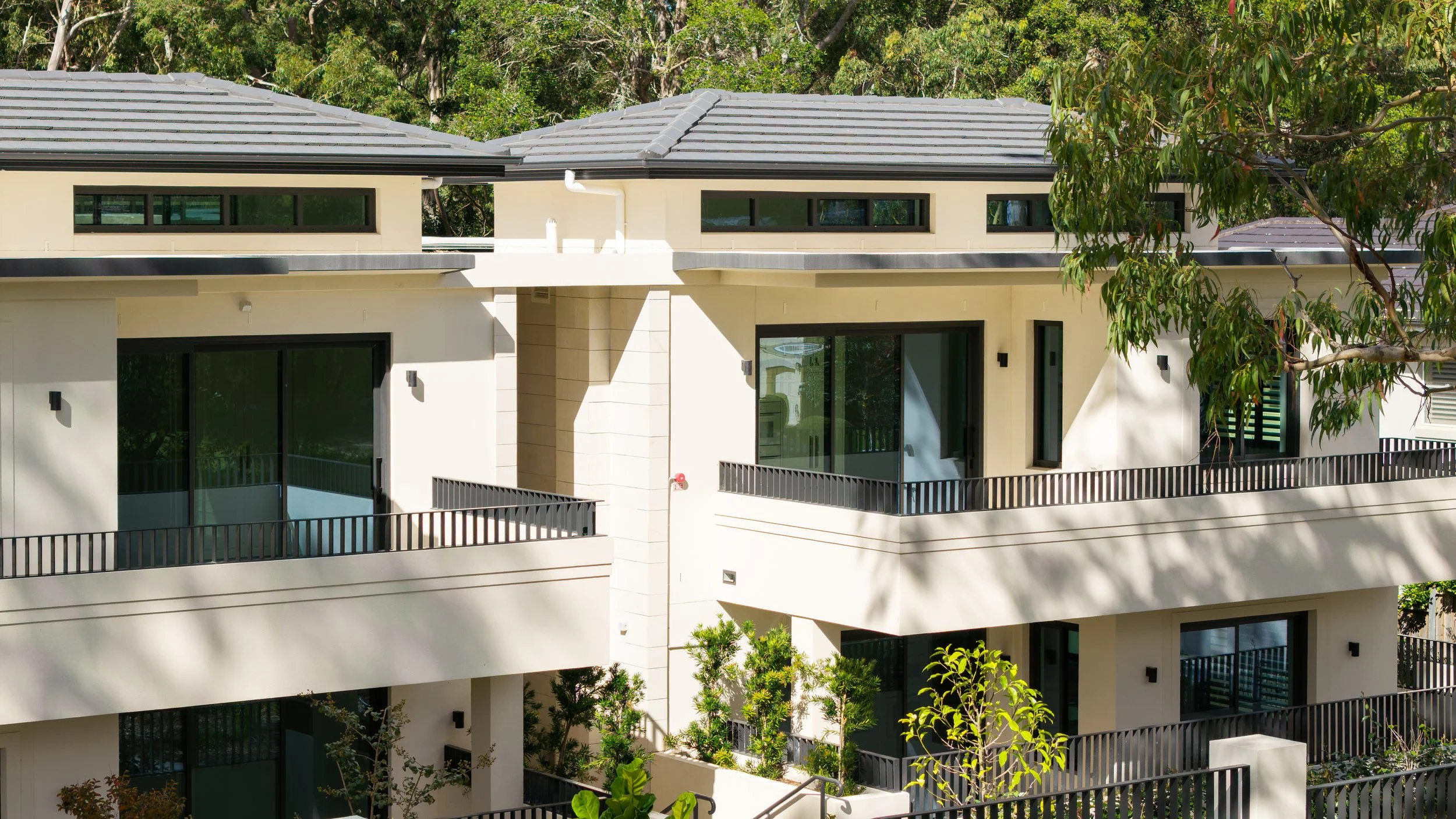 Exterior view of a modern multi-story residential building with white walls, black window frames, and balconies with black railings, surrounded by trees and greenery.