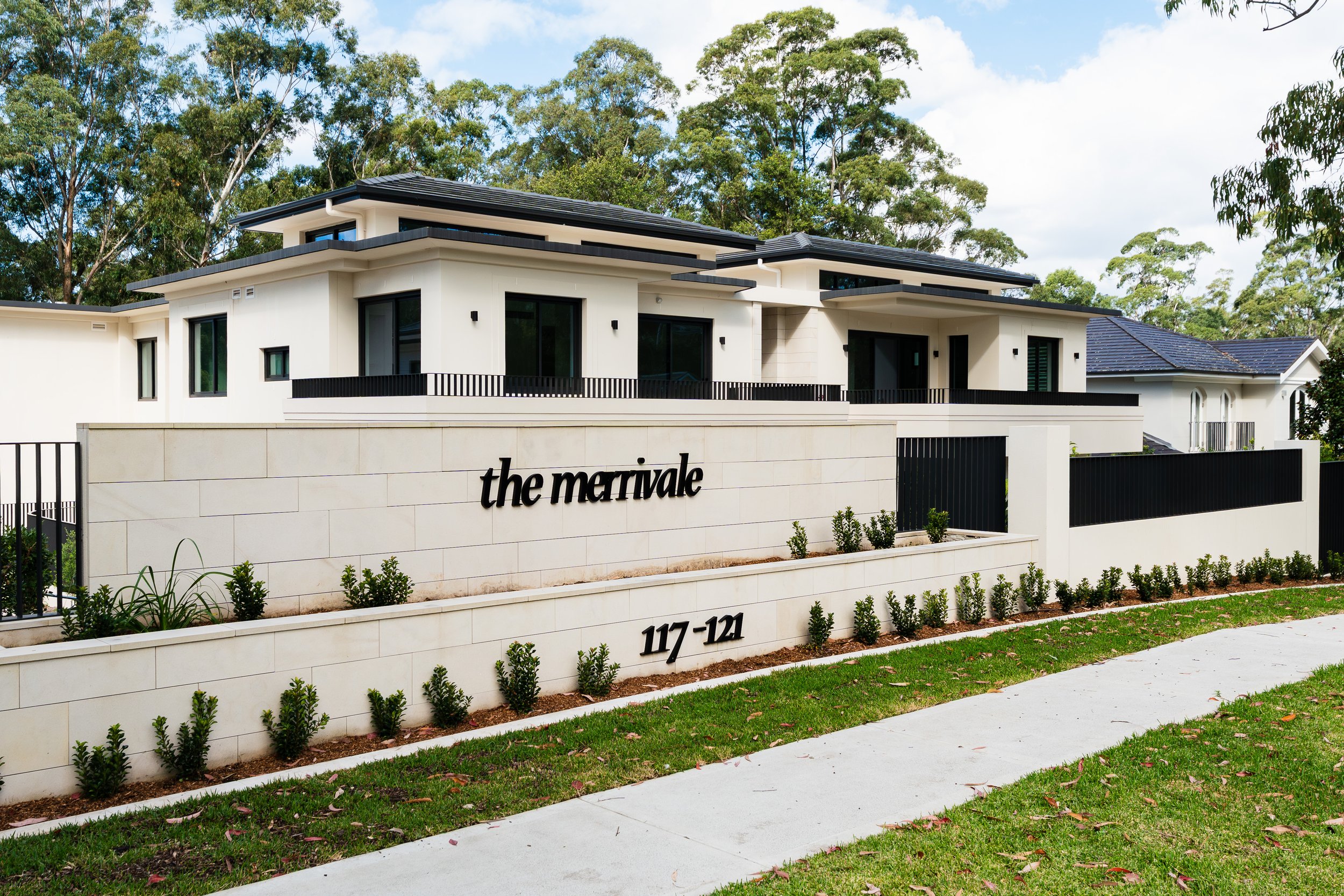 Modern multi-story residential building with white exterior walls, black window frames, and black railings, situated behind a white stone wall with the words 'the merrivale' and street number '117-121'. The building is surrounded by green trees and a