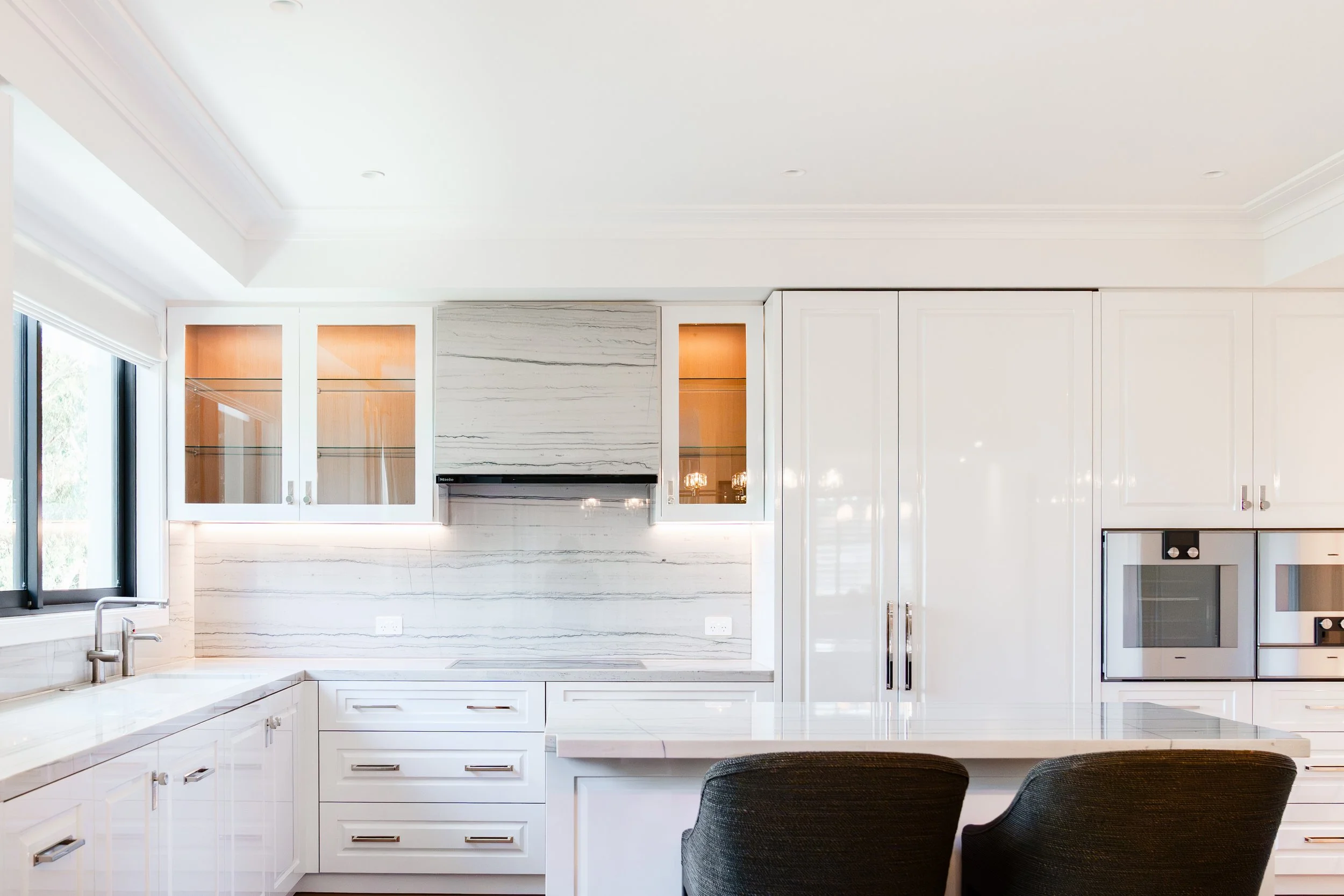 Modern white kitchen with marble backsplash, glass-front cabinets with warm lighting, built-in oven, and a kitchen island with two black chairs.