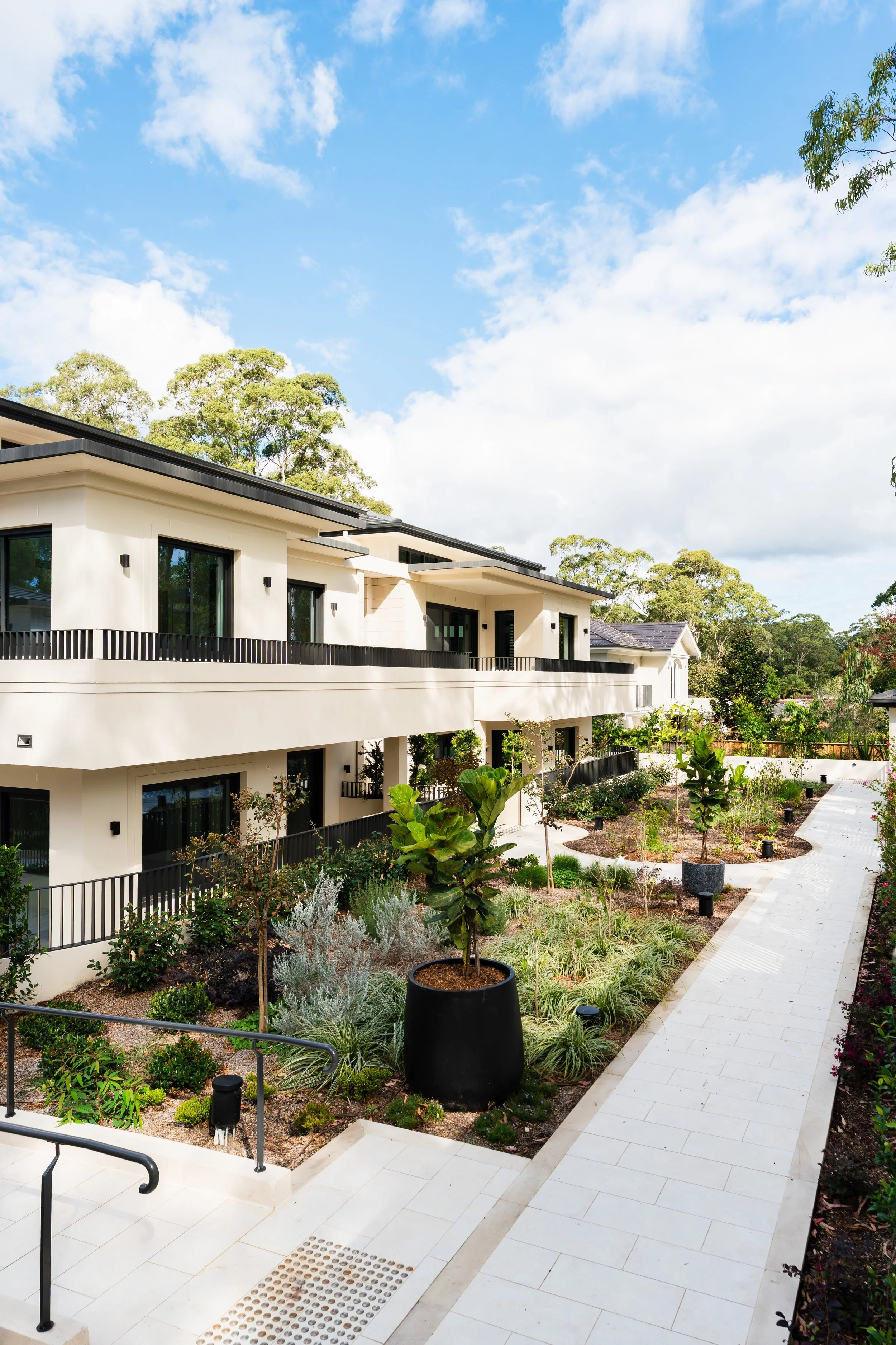 Modern multi-story white residential building with large windows, balconies, and black railings, surrounded by landscaped garden with various plants, trees, and a paved pathway under a partly cloudy sky.