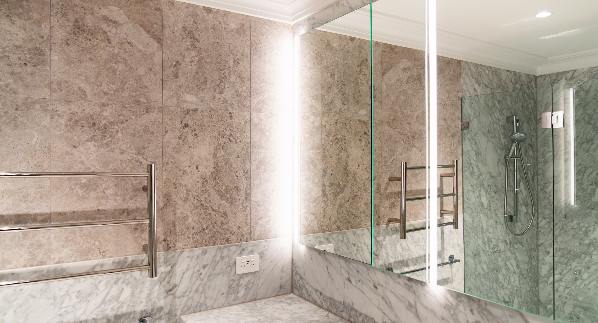 Modern bathroom with beige marble tile walls, marble floor, large mirror, glass shower enclosure with a showerhead, and a wall-mounted towel warmer.