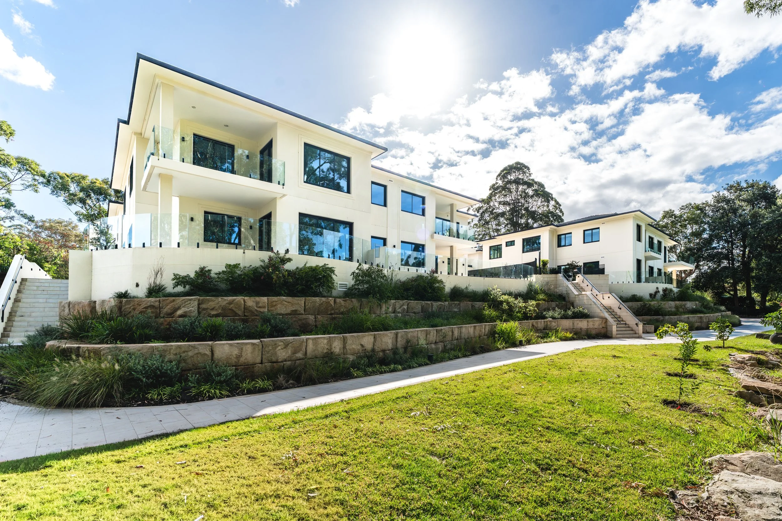 Modern white multi-story residential building with multiple balconies and large windows, surrounded by landscaped garden and stairs, under a partly cloudy sky with sunlight.