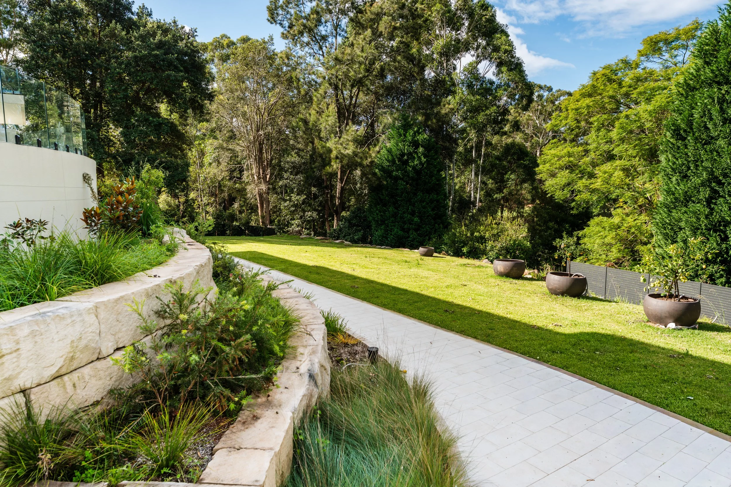 A landscaped backyard garden with a stone pathway, lush green grass, and a variety of trees and shrubs in the background on a bright, sunny day.