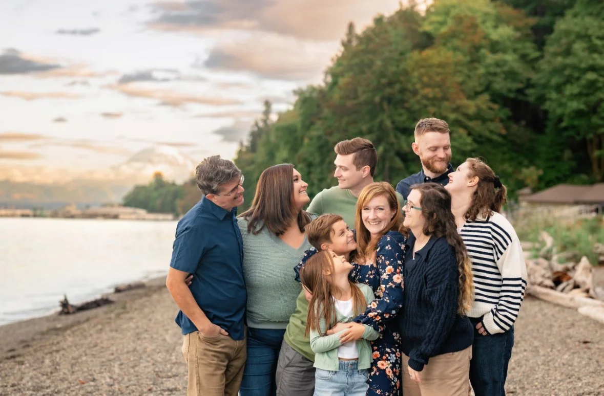 A multi-generational family standing on a beach, smiling and embracing each other, with the ocean and forested hills in the background during sunset.