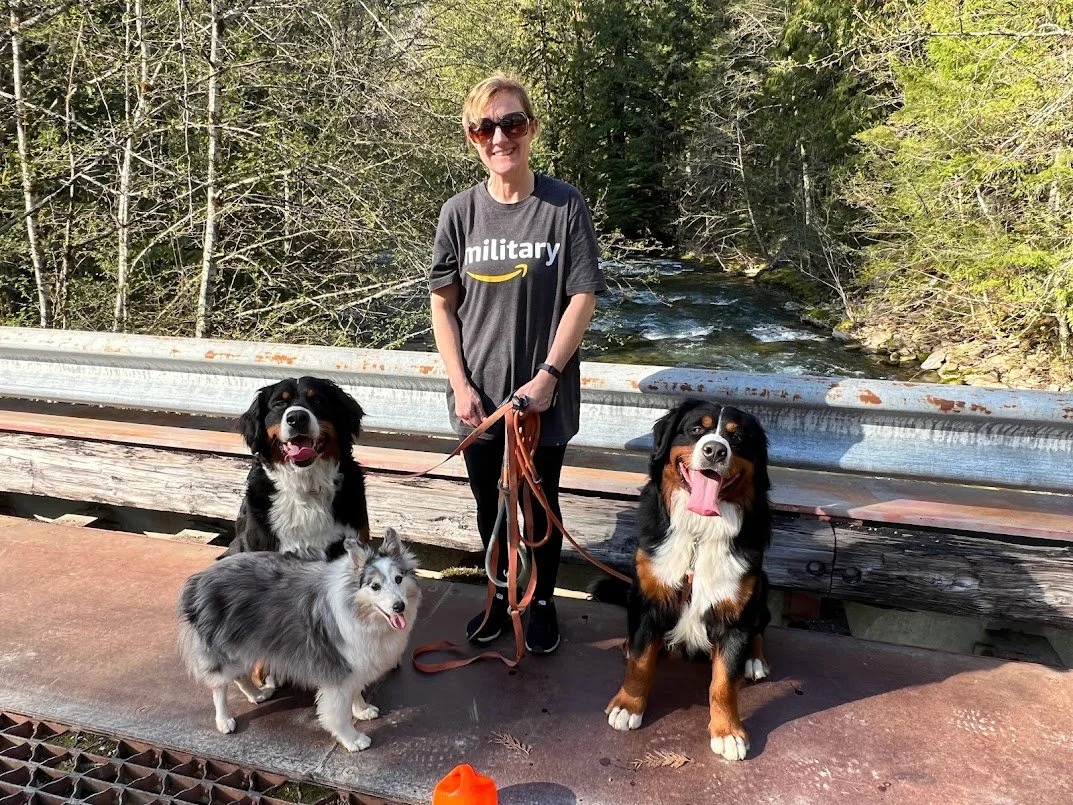 A woman smiling and wearing sunglasses, standing with three dogs on a bridge over a creek in a wooded area.
