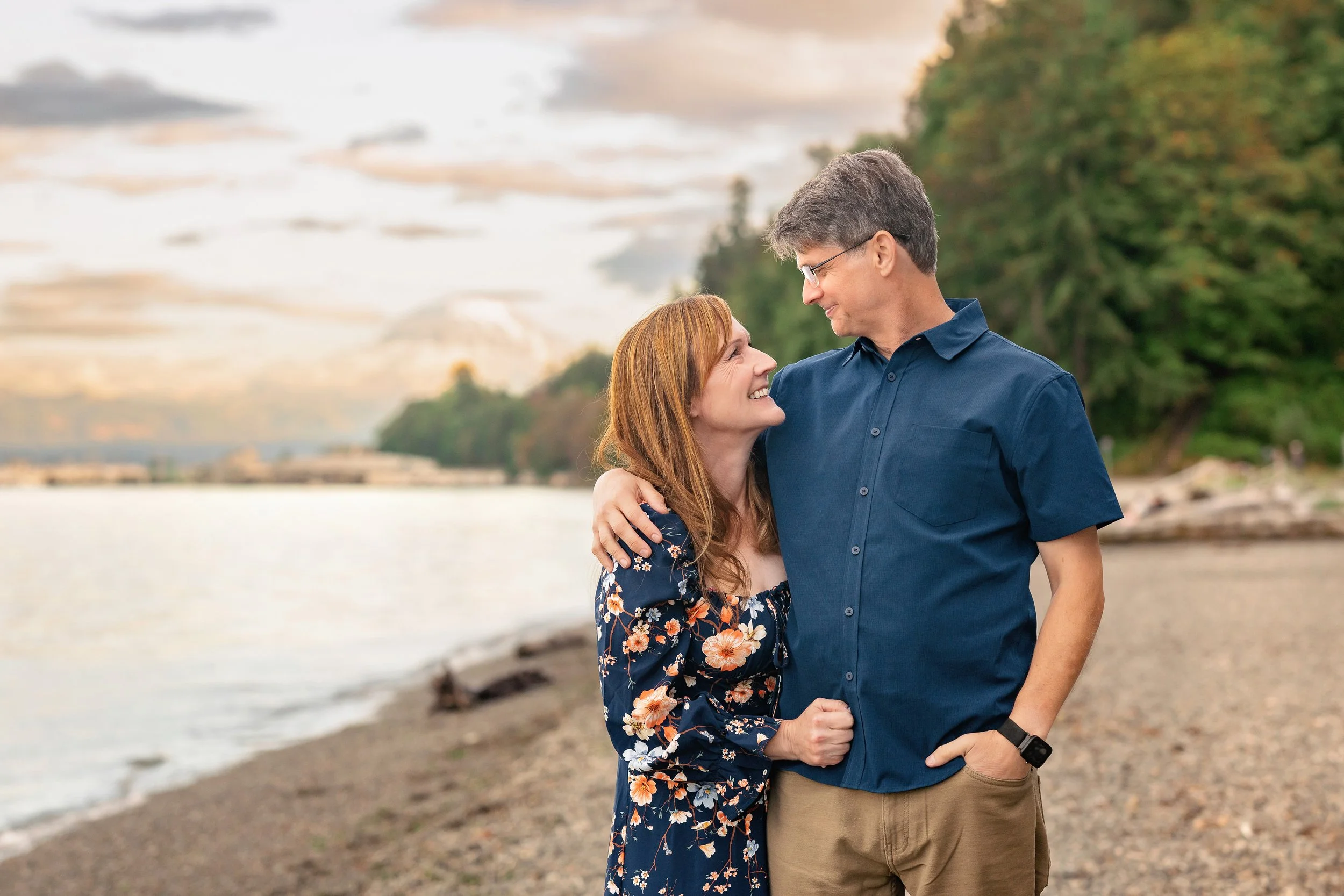 A man and woman standing close to each other on a beach, smiling and looking into each other's eyes, with water and trees in the background during sunset.