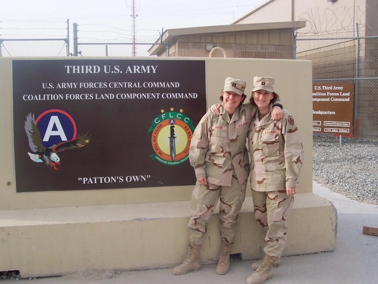 Two female soldiers in camouflage Army uniforms smiling and posing with their arms around each other in front of a sign for the Third U.S. Army at a military base.