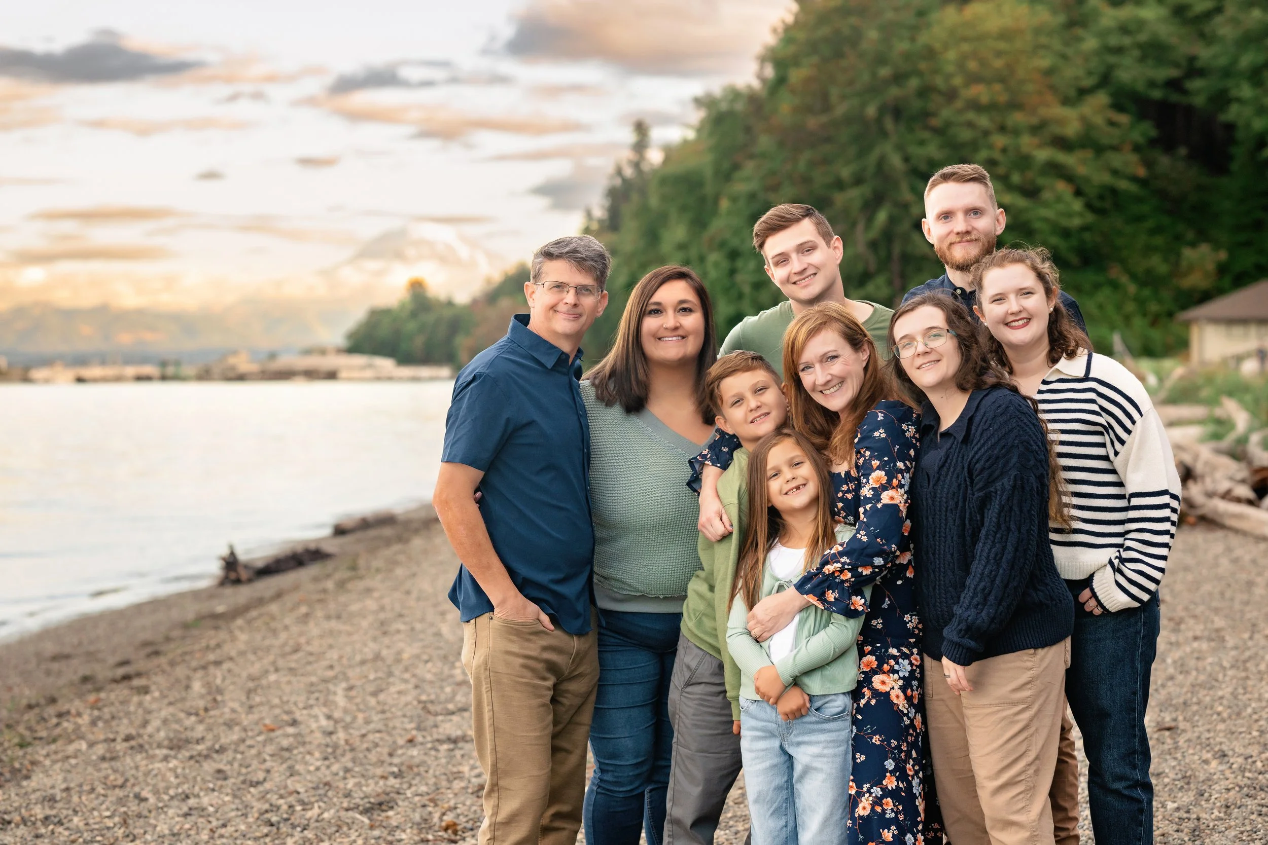 Group of nine people, including children and adults, standing together on a pebble beach near a body of water during sunset with trees and mountains in the background.