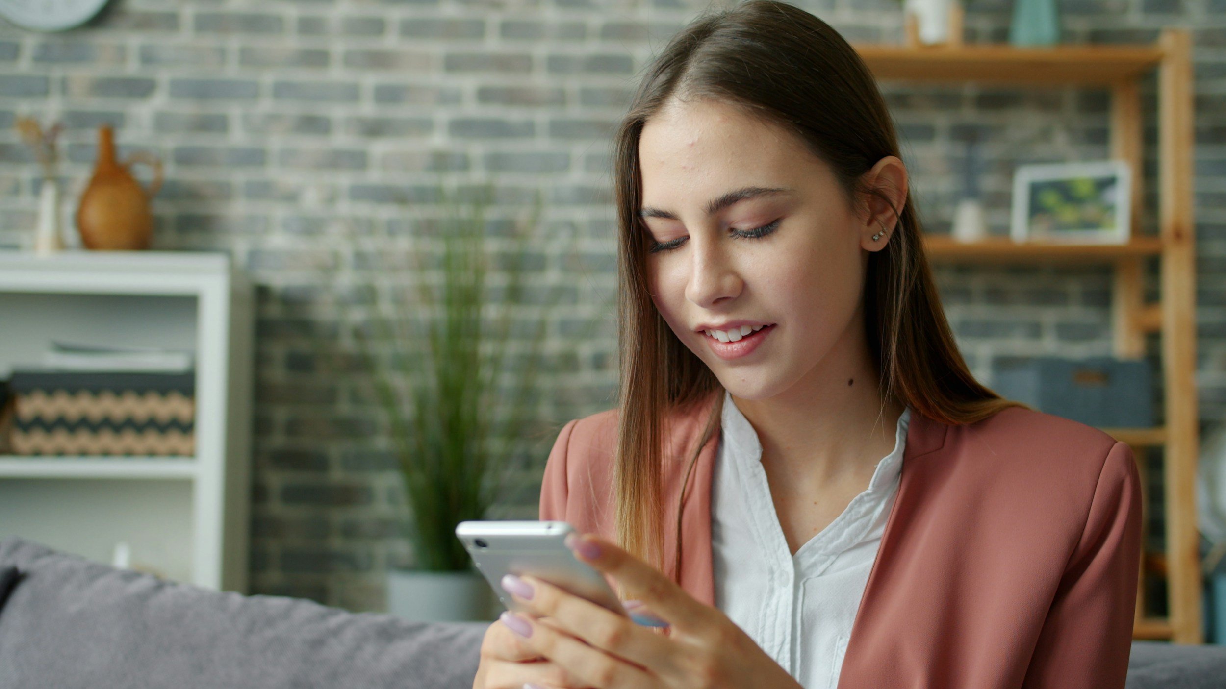 A young woman with long brown hair looking at her smartphone in a cozy room with a brick wall background and decorative shelves.