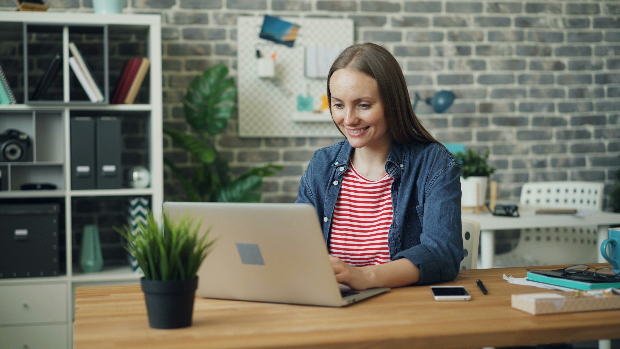 A woman with long brown hair sitting at a desk working on a laptop in a modern office with brick wall background and shelves with storage boxes and books.