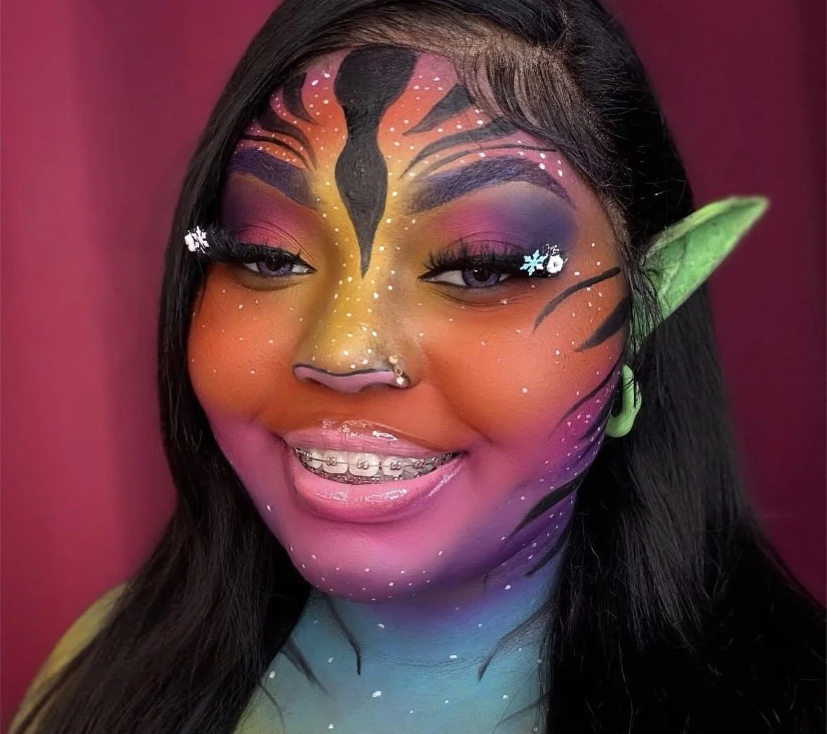 A woman with fantasy makeup resembling a dragon, featuring colorful face paint, pointed elf-like green ear, and braces.