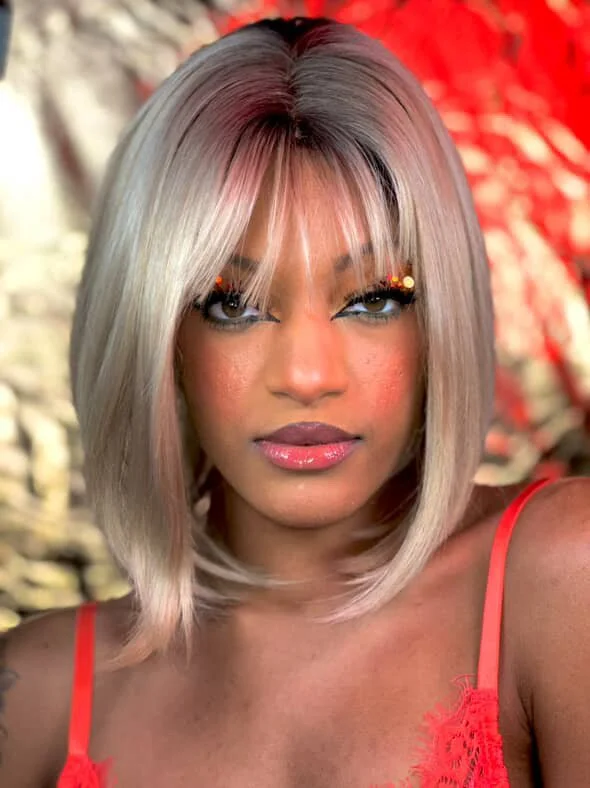 A woman with shoulder-length platinum blonde hair, makeup, and shimmering eye makeup with accents, wearing a red top with thin straps, in front of a blurred background with red and beige tones.
