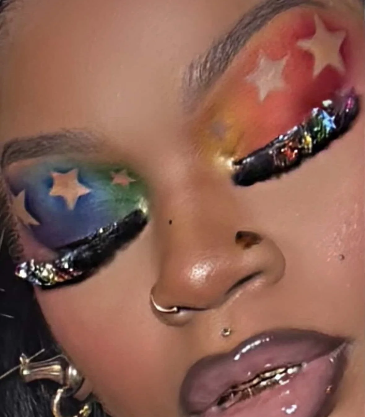 Close-up of a person's face with colorful eye makeup, glittery lips, and several facial piercings, including a nose ring, septum ring, and lip piercings.