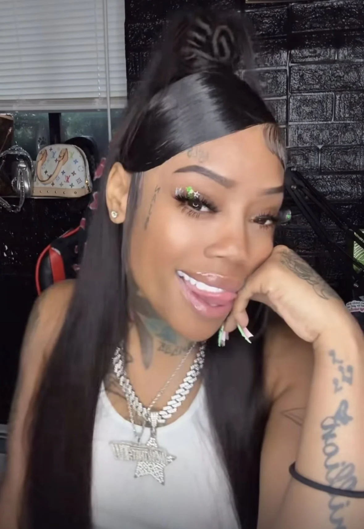 A woman with long straight black hair, smiling with her tongue slightly out, while resting her head on her hand. She is wearing makeup with glittery eye decorations, jewelry including diamond necklaces and a star pendant, and has tattoos on her arm. The background shows a black brick wall and some bags hanging on the wall.